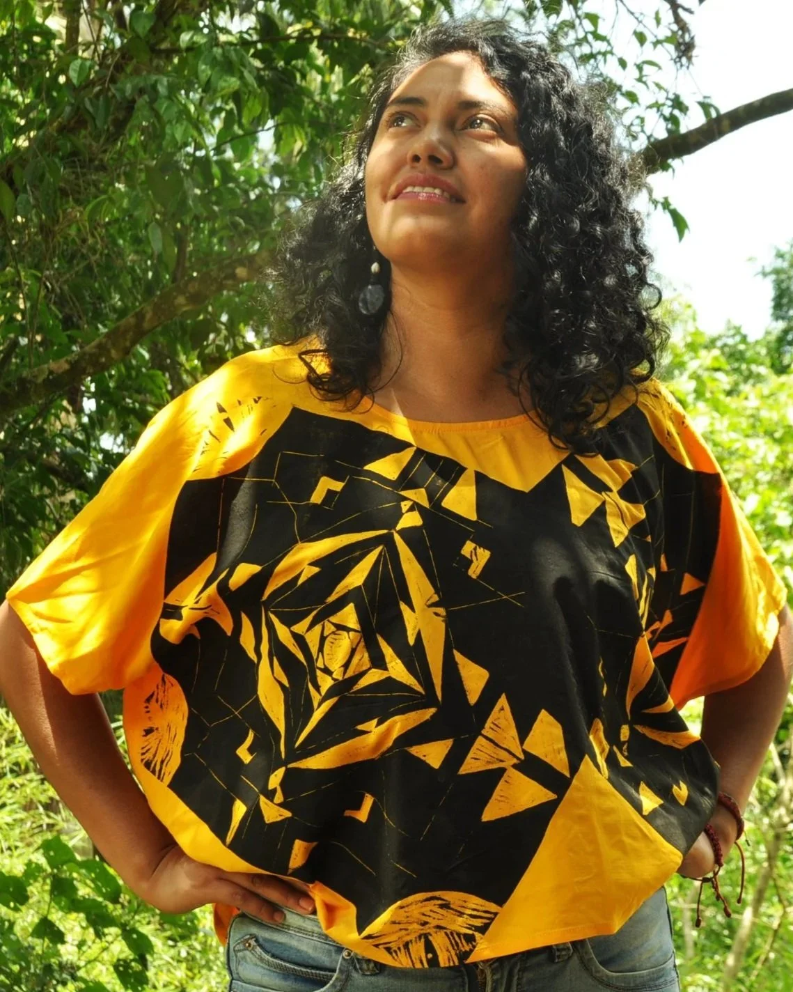 Mujer con camiseta amarilla y negra, de cabello rizado, posando en un entorno natural con árboles y vegetación.