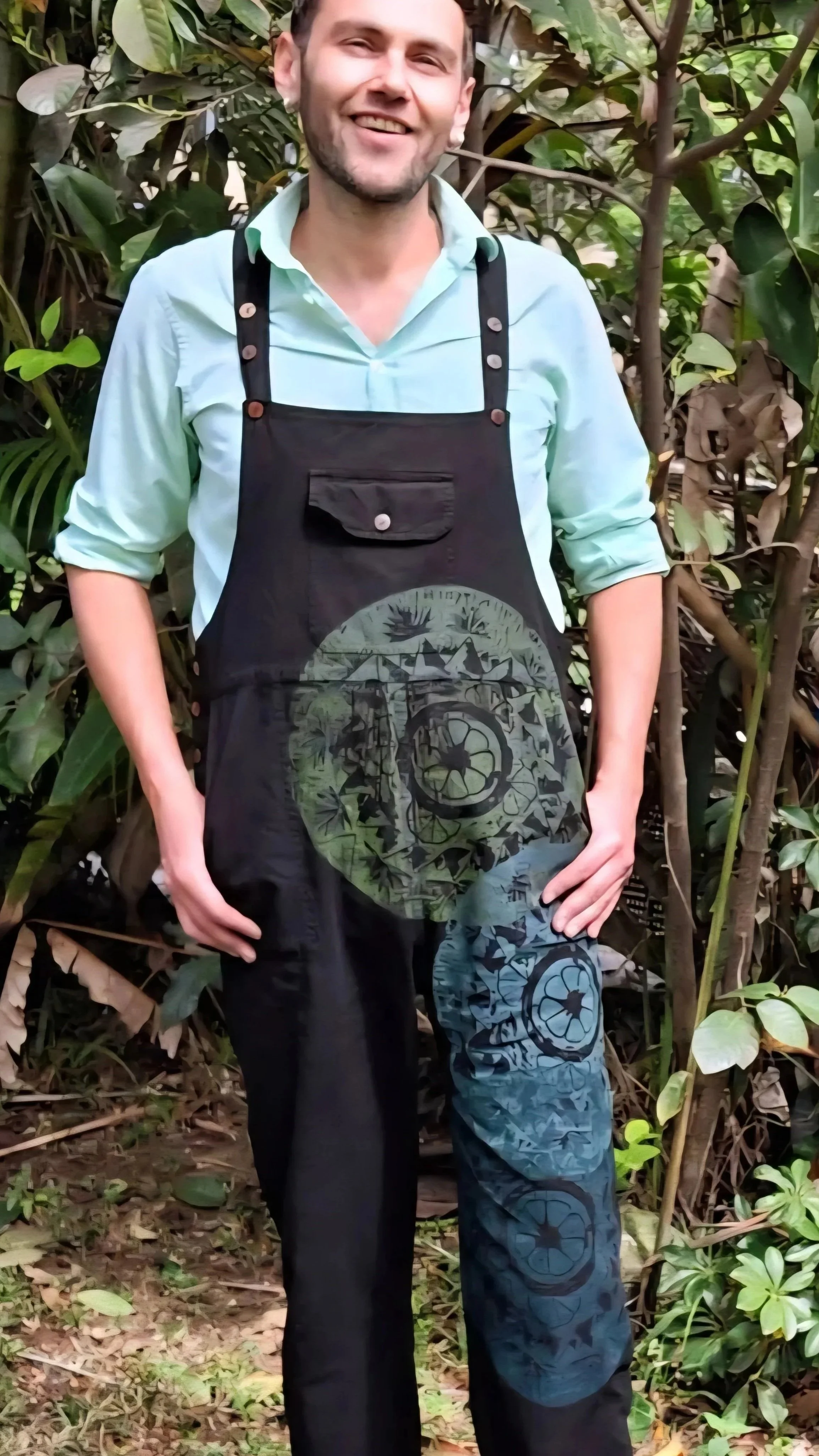 Hombre sonriendo con bigote y barba, vistiendo una camisa de color menta con la manga enrollada y un delantal negro con un diseño artístico, en un entorno de vegetación densa.