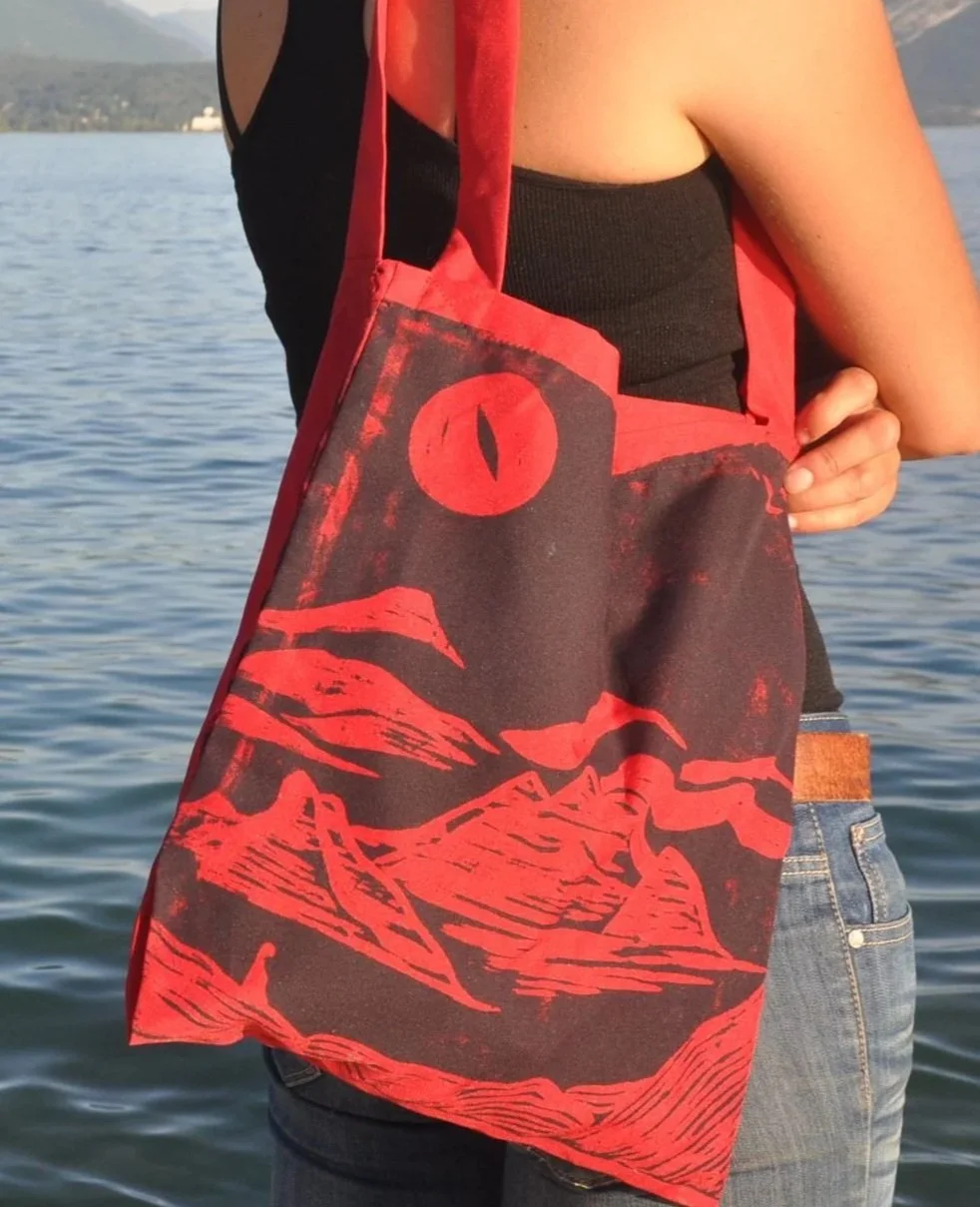 Persona con camiseta negra y pantalones vaqueros, sosteniendo una bolsa de tela roja y negra con diseño de montañas y sol, junto a un cuerpo de agua.