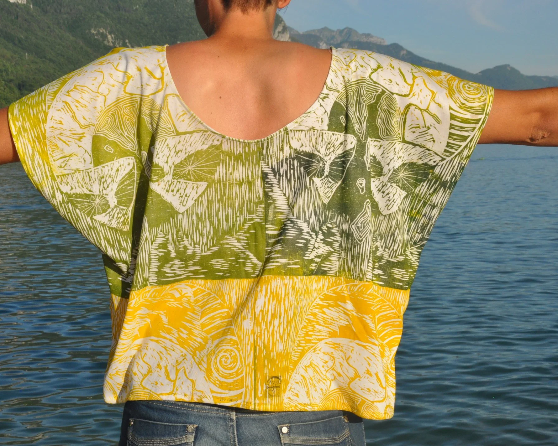 Persona de espaldas con brazos extendidos en un cuerpo de agua con montañas al fondo, usando una blusa suelta con un diseño colorido en tonos verdes y amarillos.