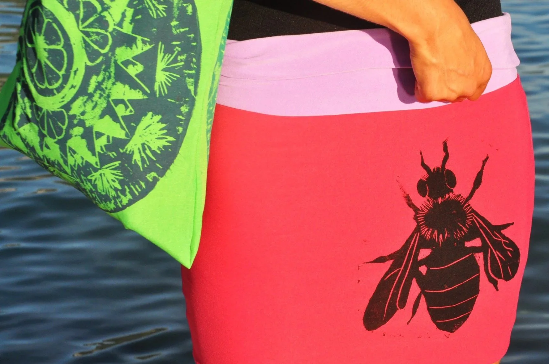 Persona con ropa de colores brillantes, mostrando un pantalón rosa con una impresión de abeja negra, una camisa verde con un diseño de frutas de limón. Fondo de agua.