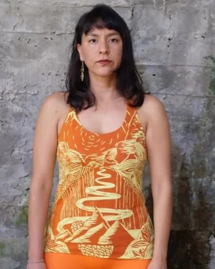 Mujer con cabello oscuro y flequillo, usando un vestido sin mangas de color naranja con diseño amarillo sobre un fondo de muro de madera.