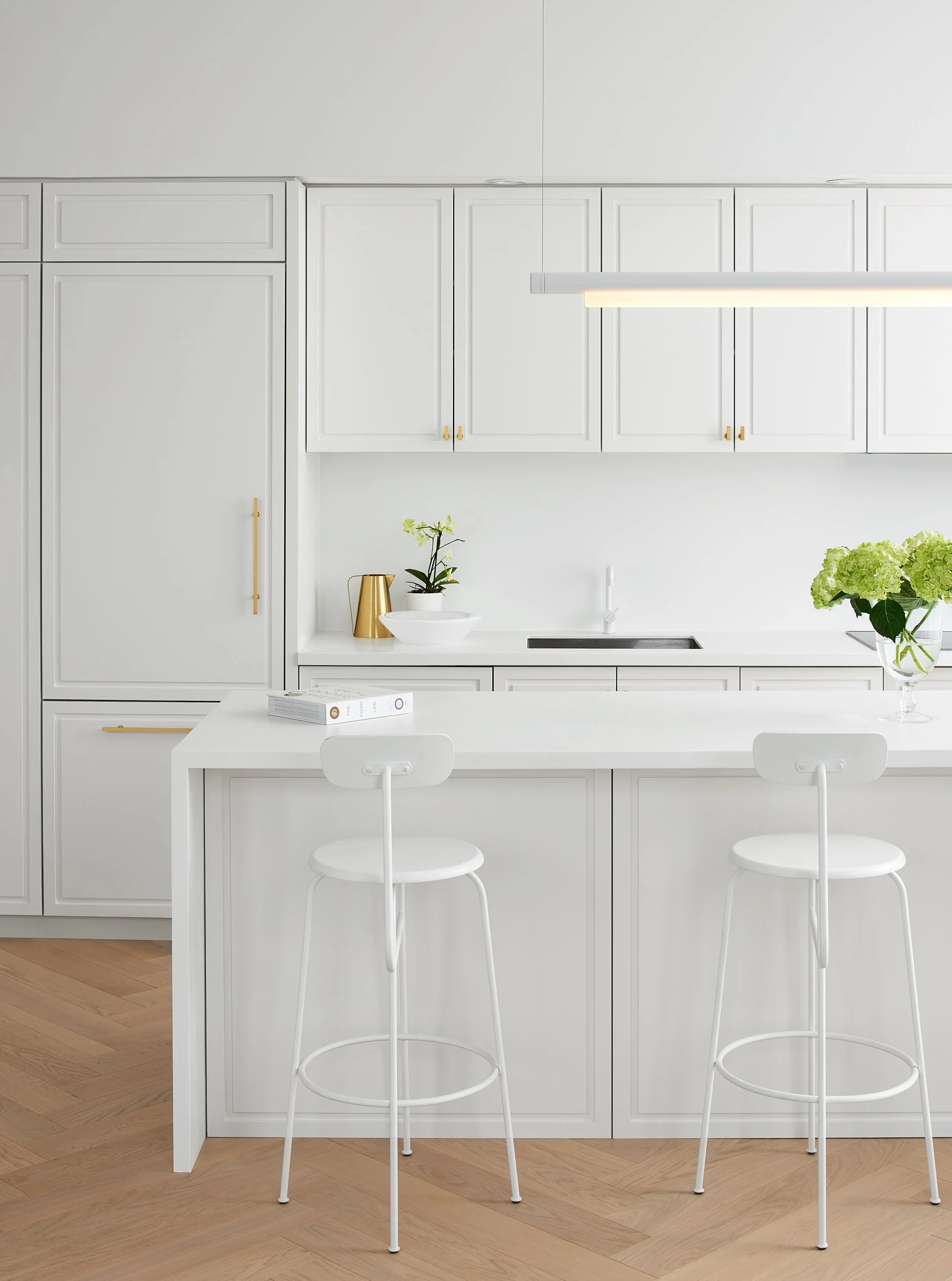 Modern white kitchen with cabinets, island, bar stools, plants, and gold accents.