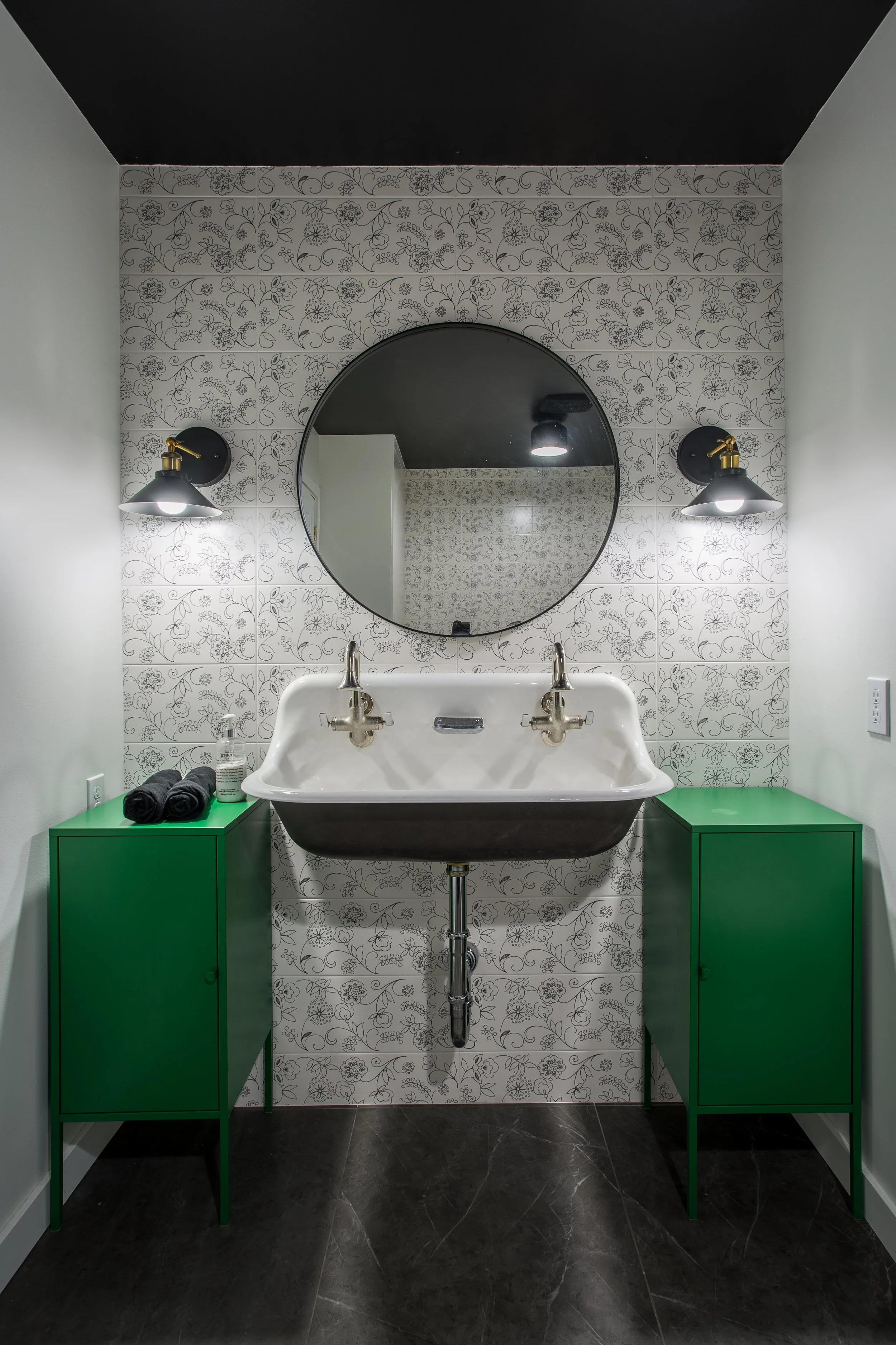 A bathroom with a white rustic sink, black piping, a round mirror, two black and brass wall sconces, green cabinets on each side, black painted ceiling, and black tiled floor.