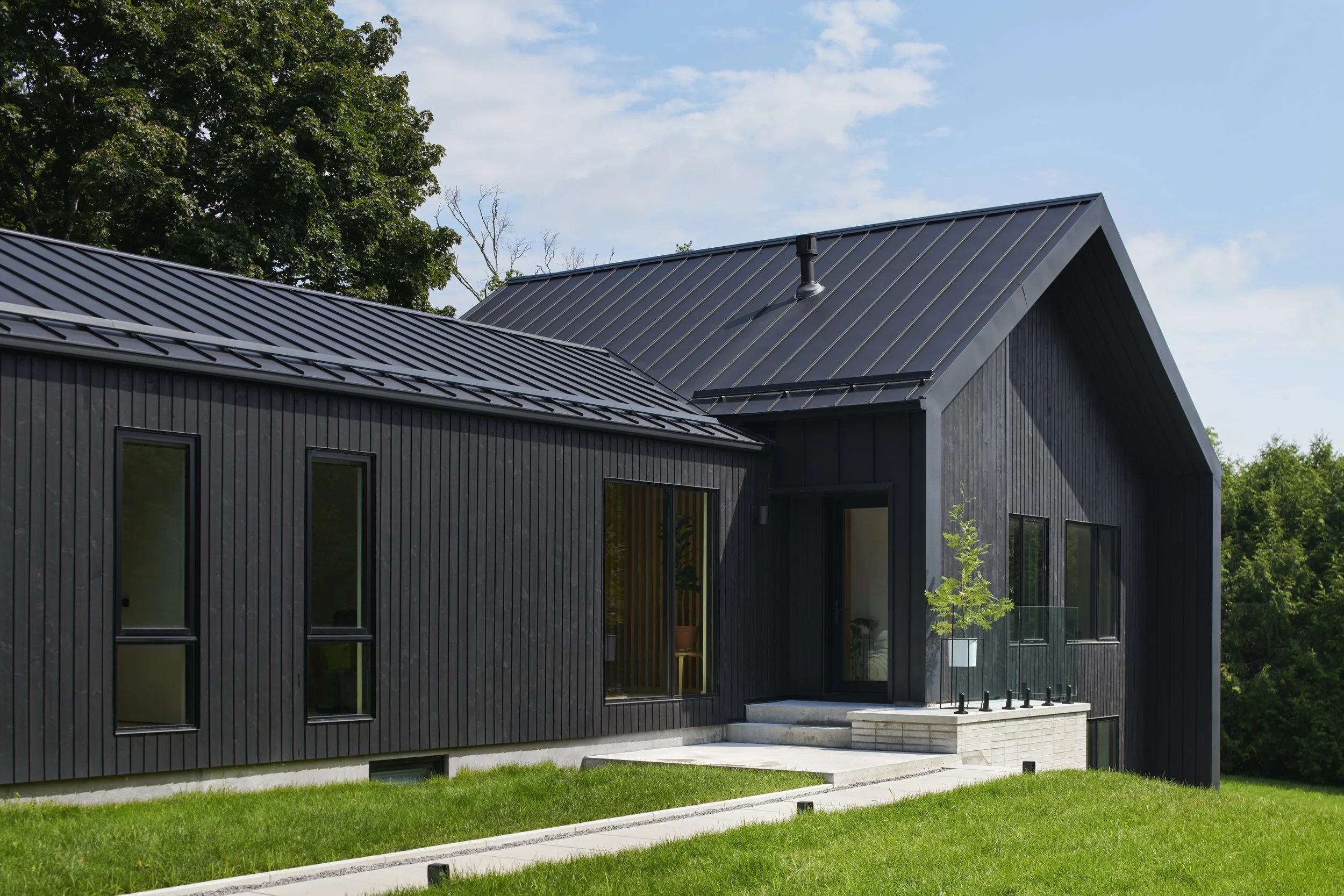 Moder rennovation with black wooden siding, metal roof, and front steps leading to the entrance, surrounded by green lawn and trees in the background under a partly cloudy sky.
