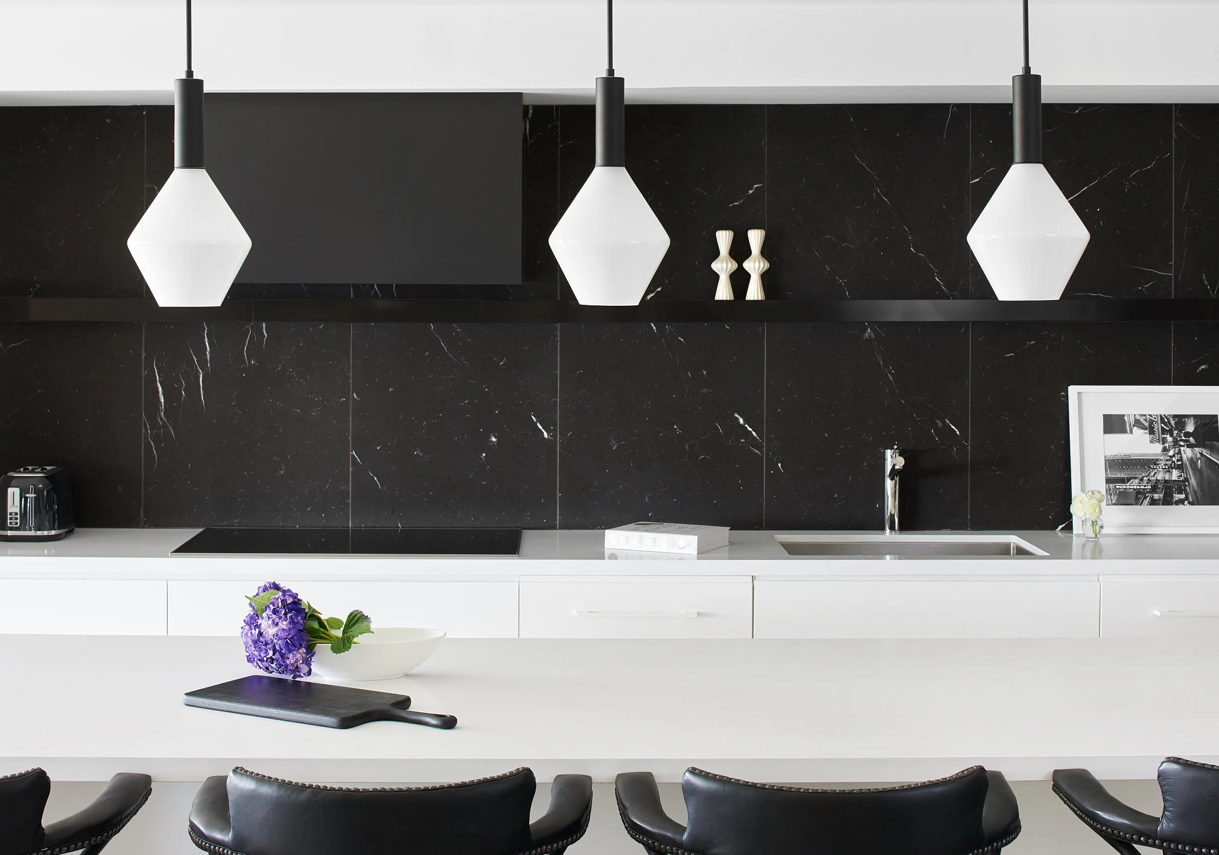 Modern black and white kitchen with pendant lights, black marble backsplash, white counter, and black chairs.