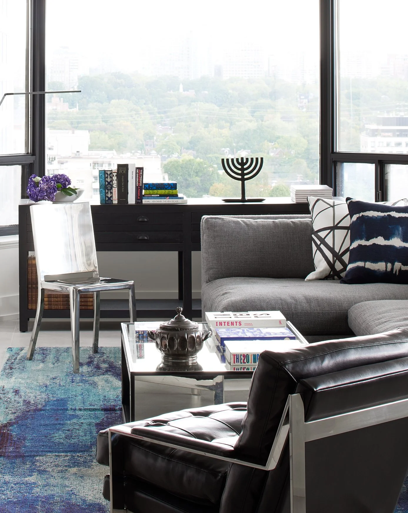 Modern living room with large windows, black console table with books, a silver menorah, and decorative items; gray sofa with patterned pillows; black leather chair; glass coffee table with books and a decorative artifact; blue patterned rug.