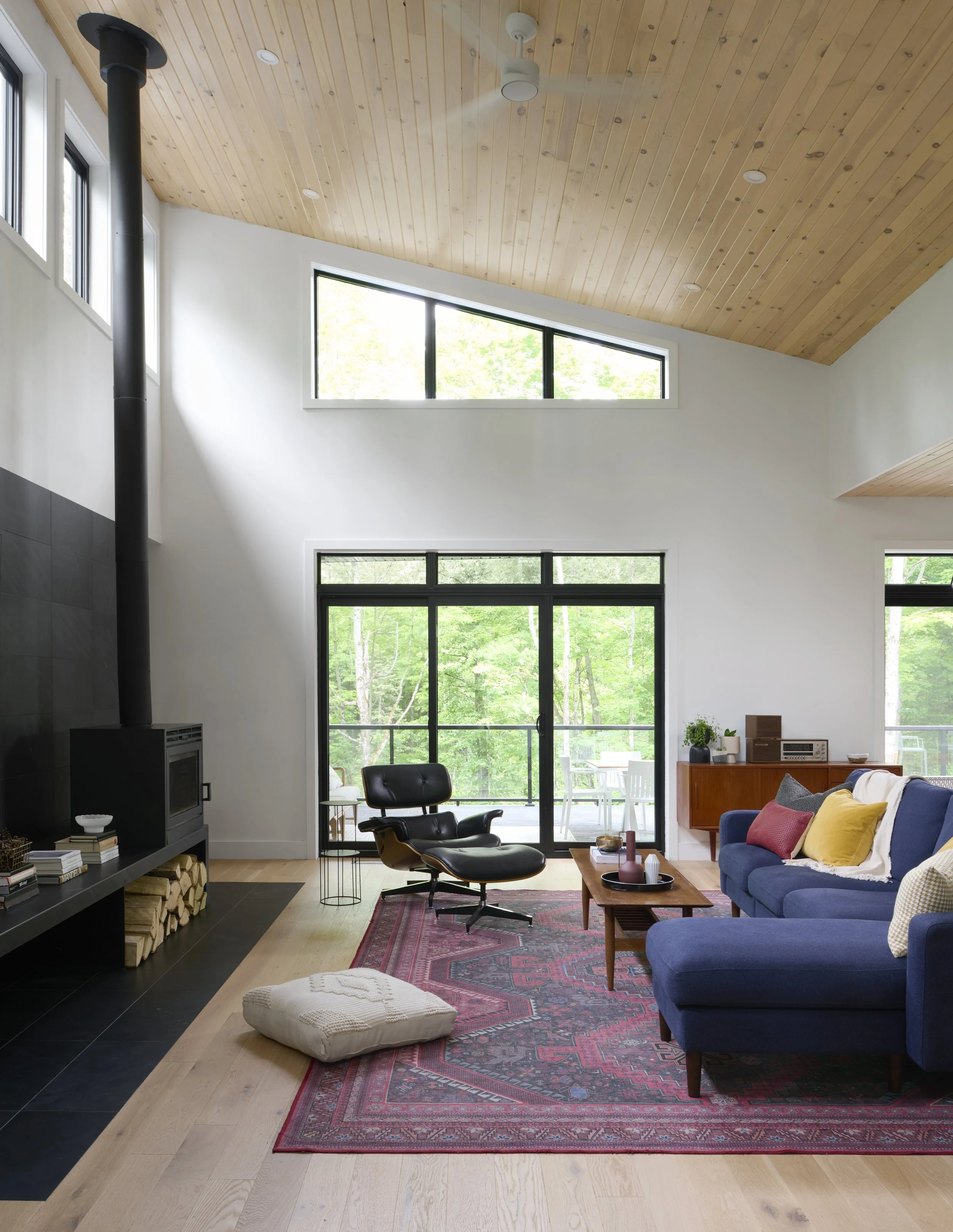 A modern living room with a high, wooden ceiling, large windows, a blue sofa with colorful pillows, a black lounge chair, a red patterned rug, a fireplace with wood stacked underneath, and a sliding glass door leading to an outside deck with trees.