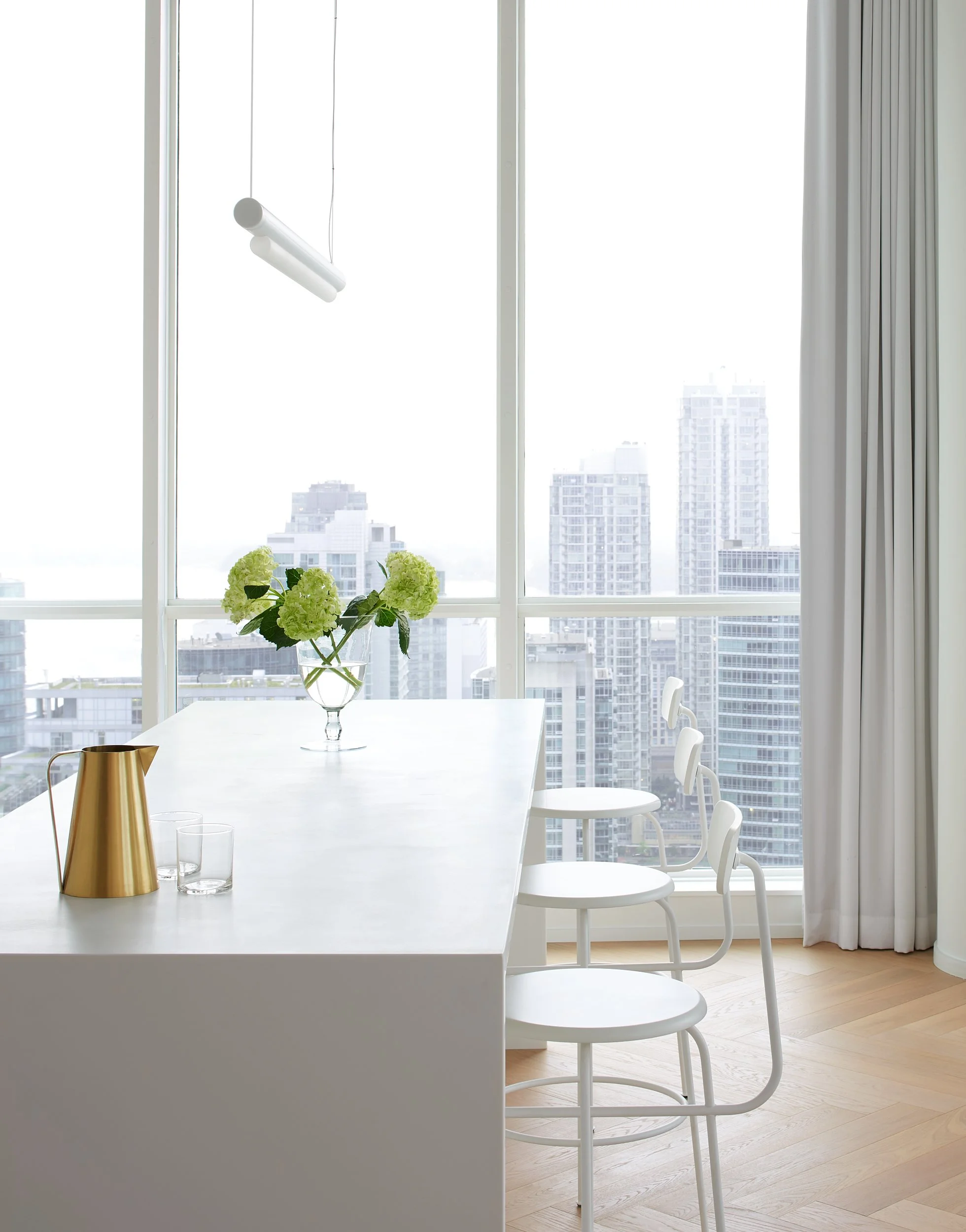 Modern dining area with a white table, a floral centerpiece, a gold pitcher, three glasses, and three white chairs in front of large windows with a city skyline.