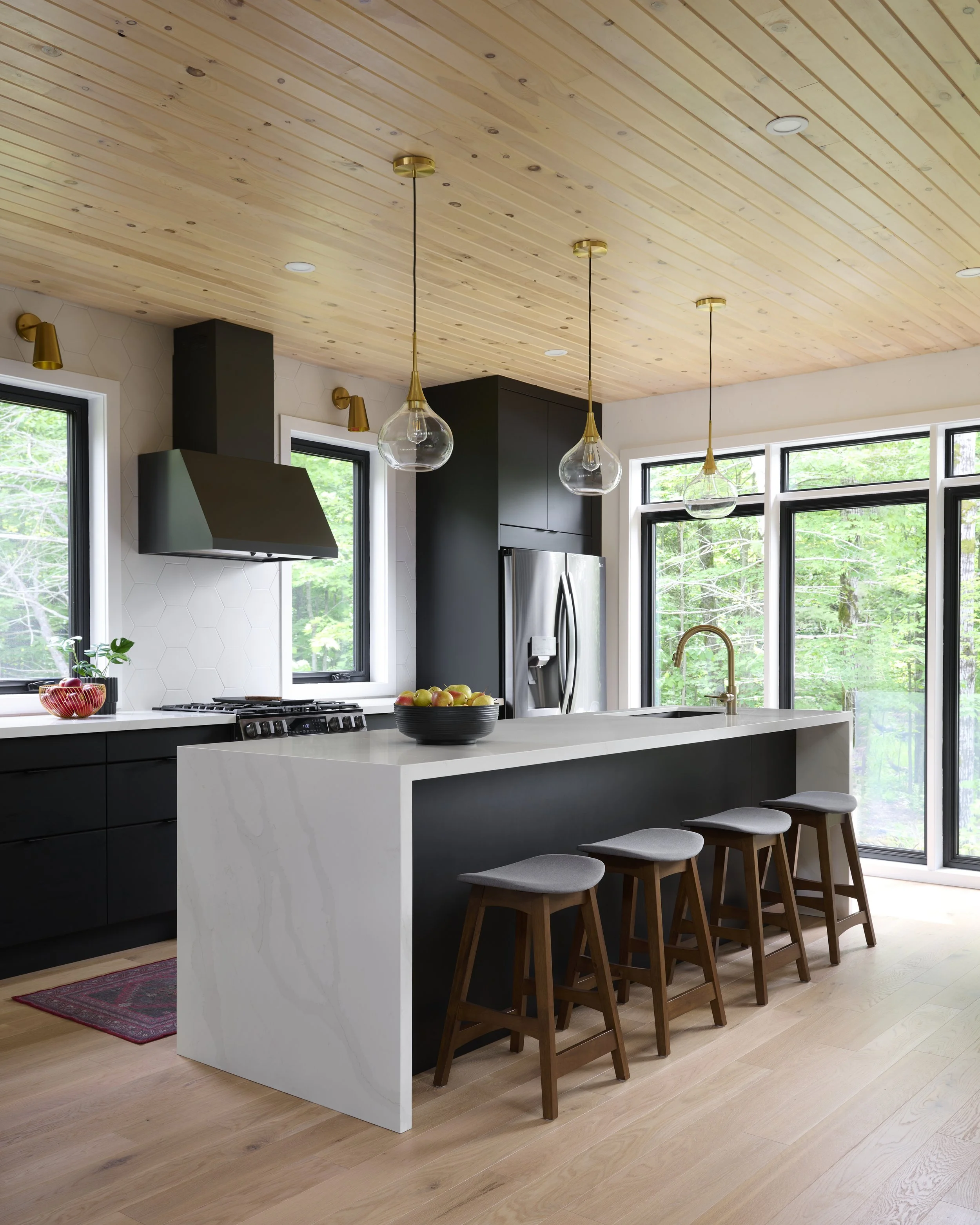 Modern kitchen with black cabinets, large windows, a white marble island with a black accent, four wooden stools with gray cushions, and pendant lights hanging from a wooden ceiling.