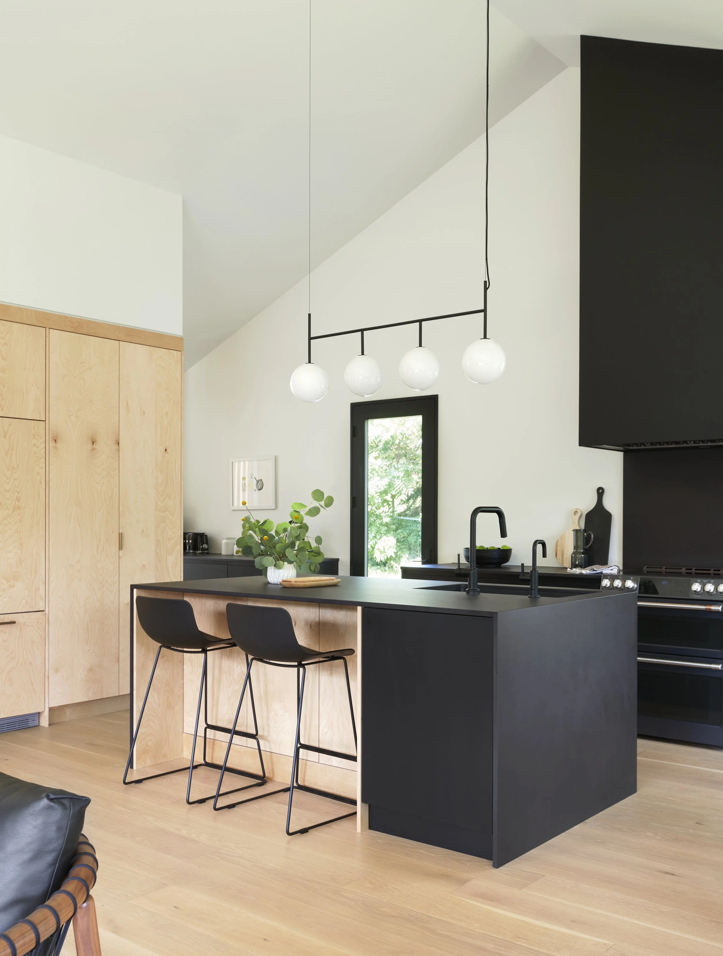 Modern kitchen with black island, two black bar stools, wooden cabinetry, hanging light fixture with four white globes, black window frame, black appliances, and minimal decor.
