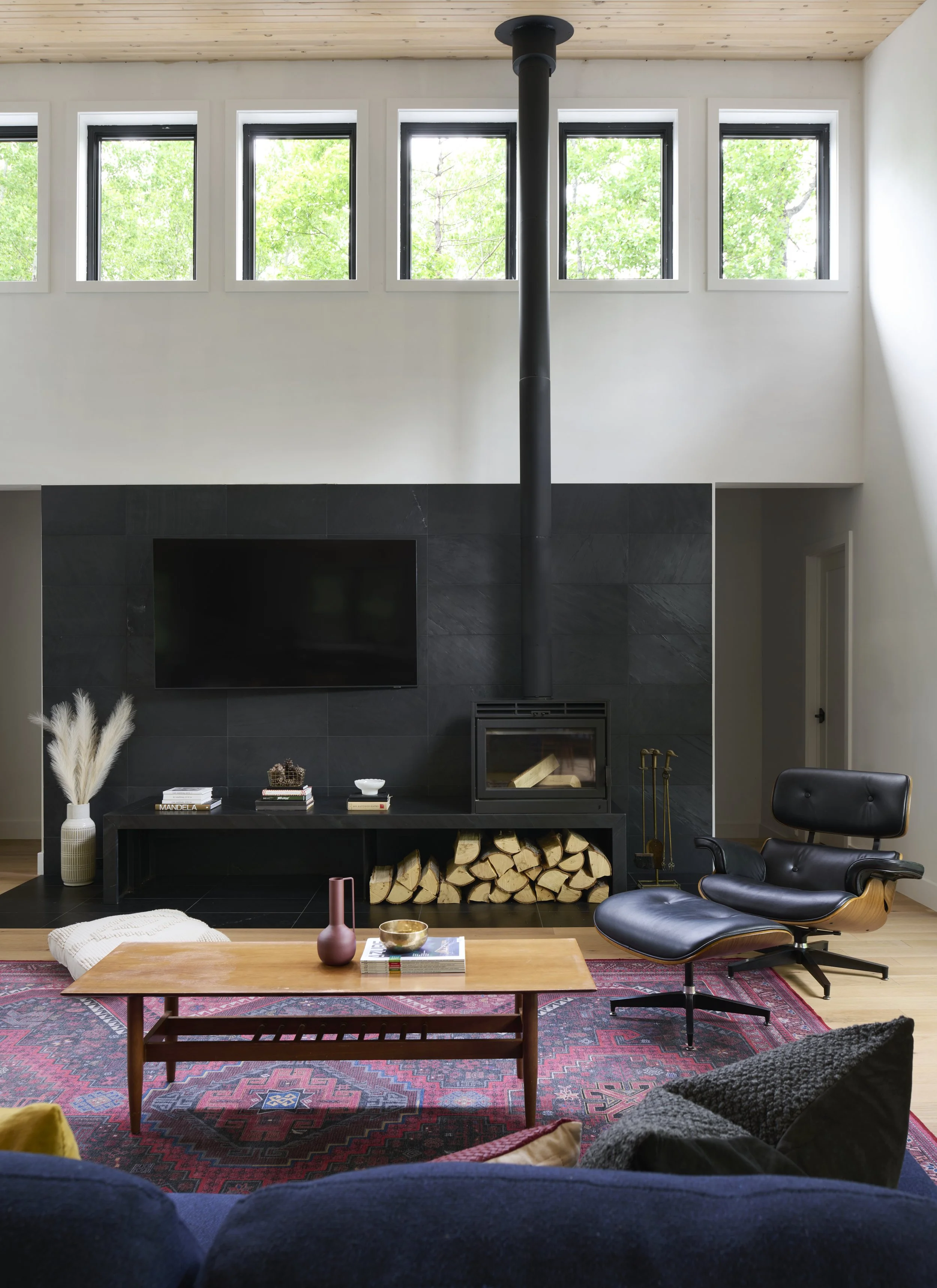 Modern living room with a black fireplace, a mounted flat-screen TV, logs stored underneath, a black lounge chair with ottoman, a wooden coffee table with decorative vases and books, a patterned area rug, and large windows with greenery outside.