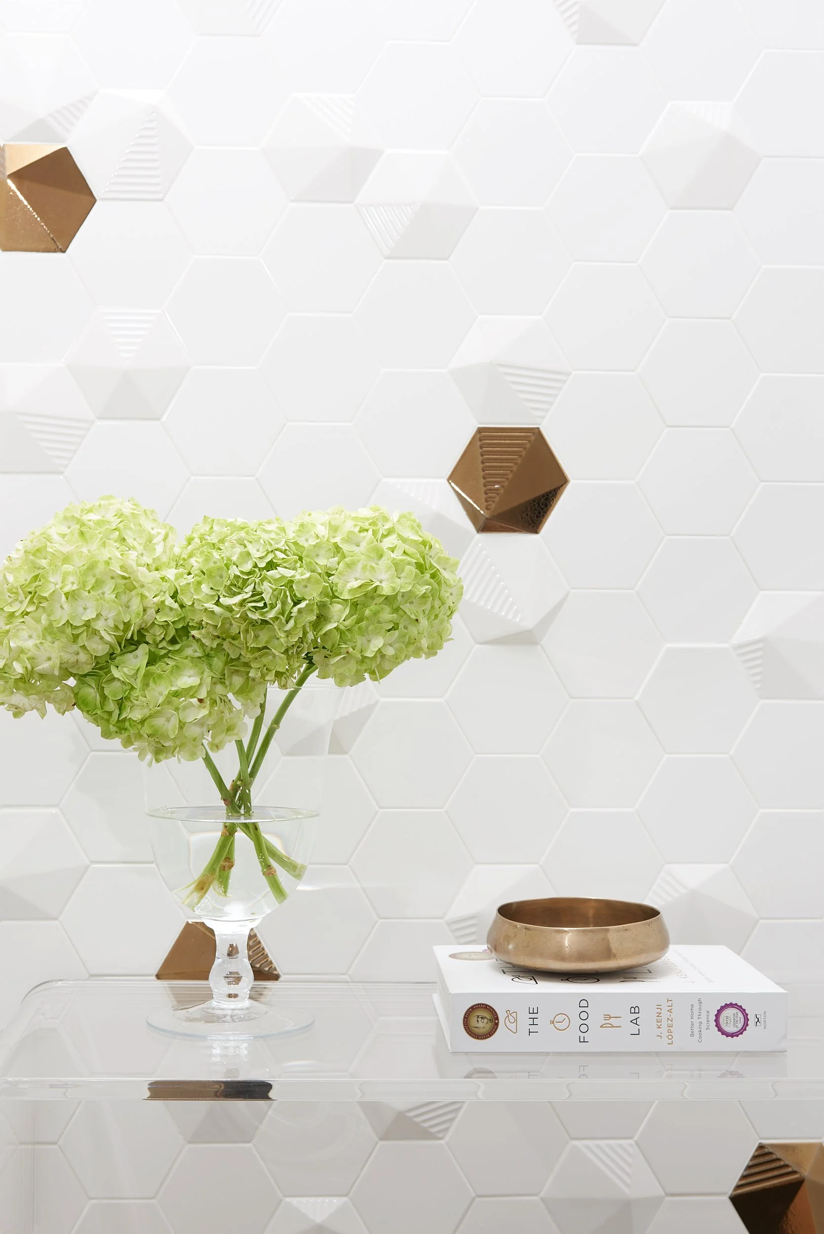 A clear glass vase with green hydrangea flowers on a transparent surface, with a brass bowl and a cookbook titled 'The Food Lab' on top, against a white hexagonal tiled wall with some brown and textured tiles.