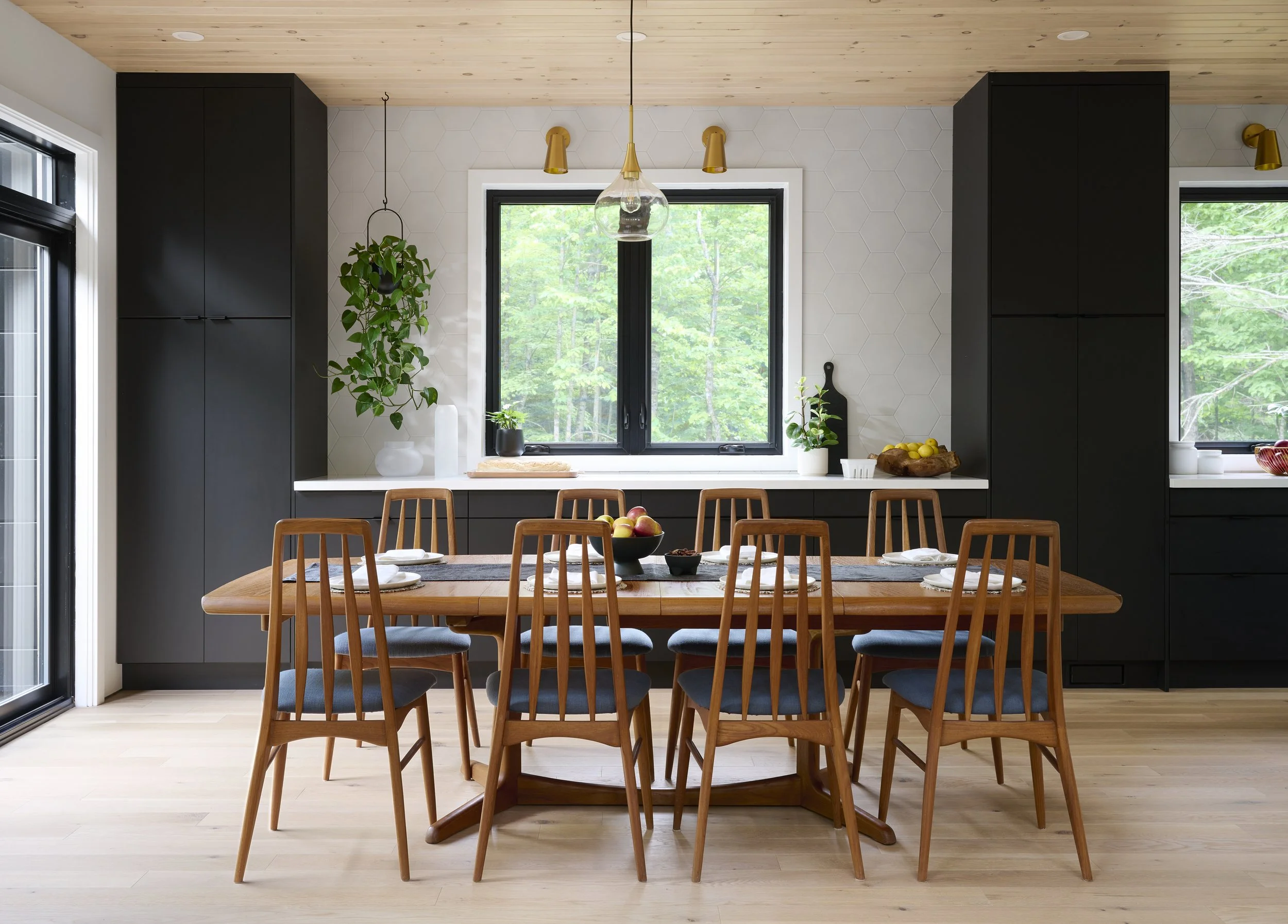 Modern kitchen with a wooden dining table set with chairs, surrounded by black cabinetry, a window showing greenery outside, and minimal decor including plants and bowls of fruit.