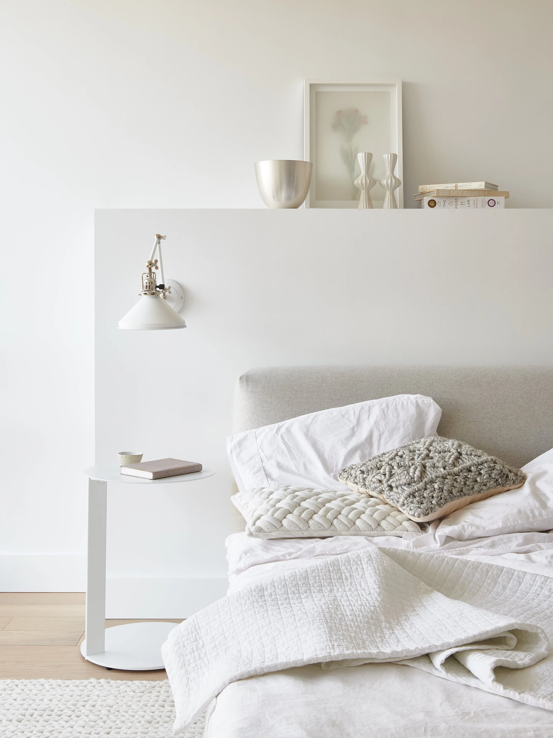 Minimalist bedroom with white bedding, decorative pillows, a side table with a book and a small bowl, a wall-mounted lamp, and a white shelf with vases and art.