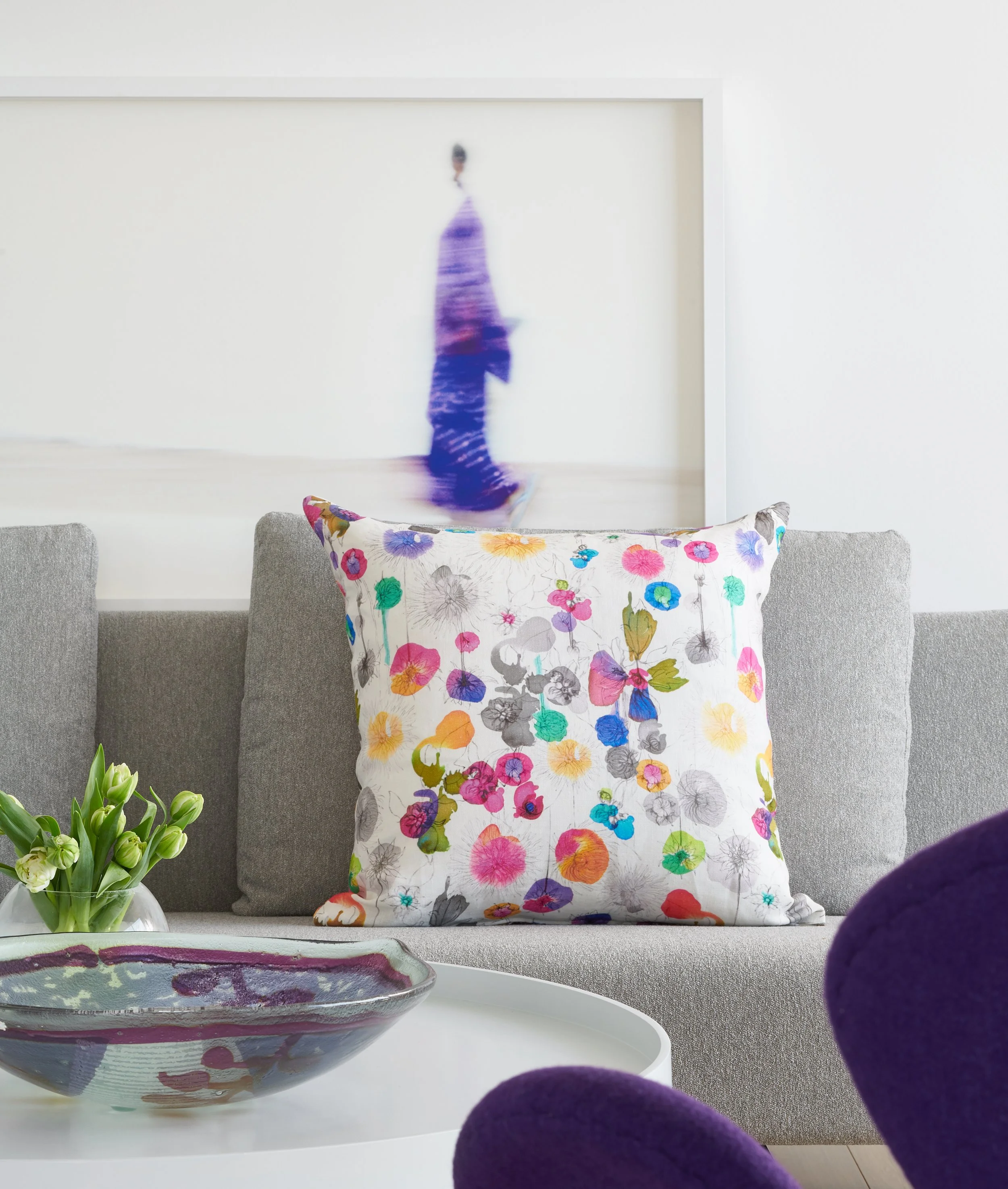 A colorful floral-patterned pillow on a gray sofa in front of a large abstract painting and a glass bowl with flowers on a white table.