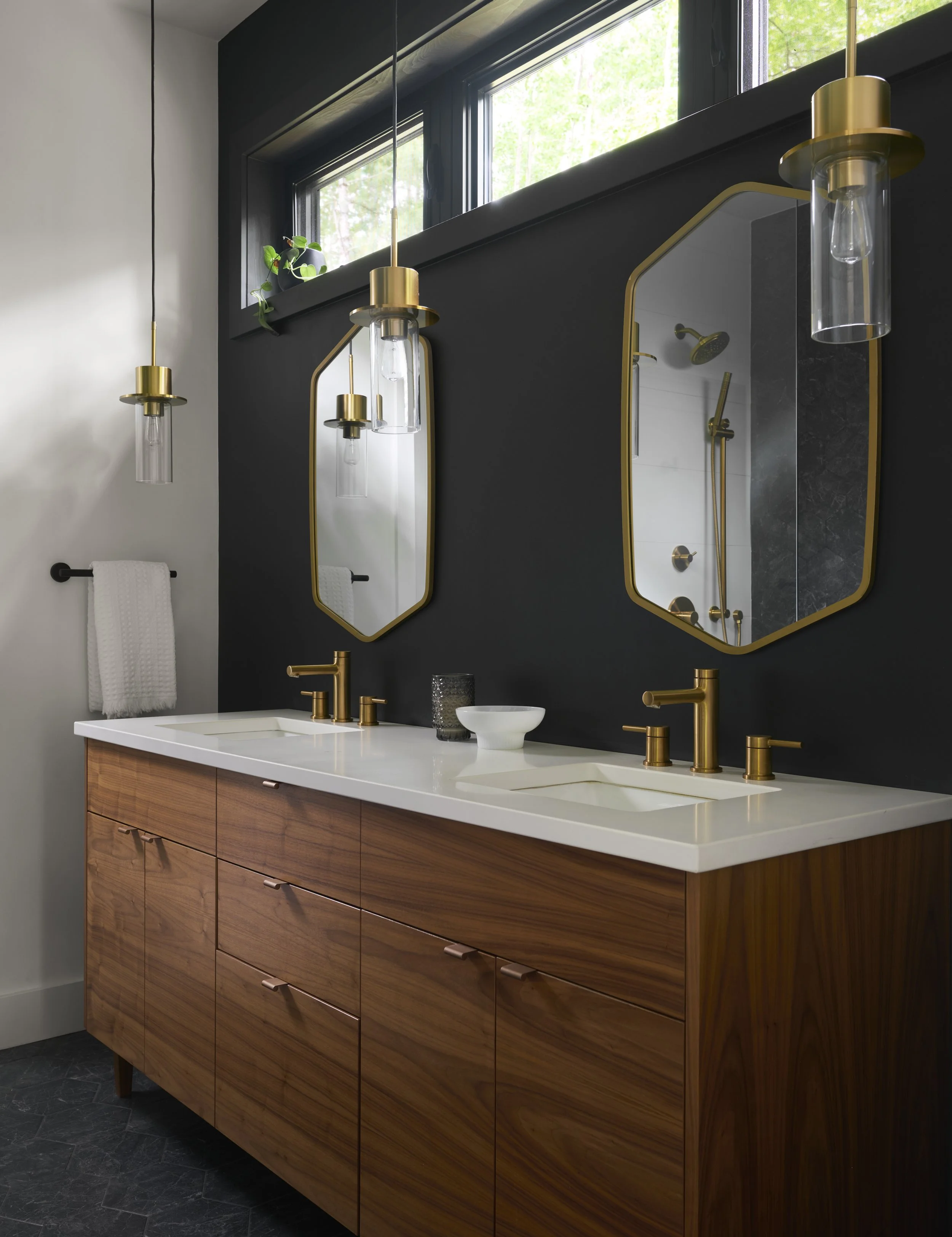 Modern bathroom with a black accent wall, gold fixtures, two vertically-oriented mirrors with gold frames, wooden vanity with a white countertop, and pendant lights hanging from the ceiling.