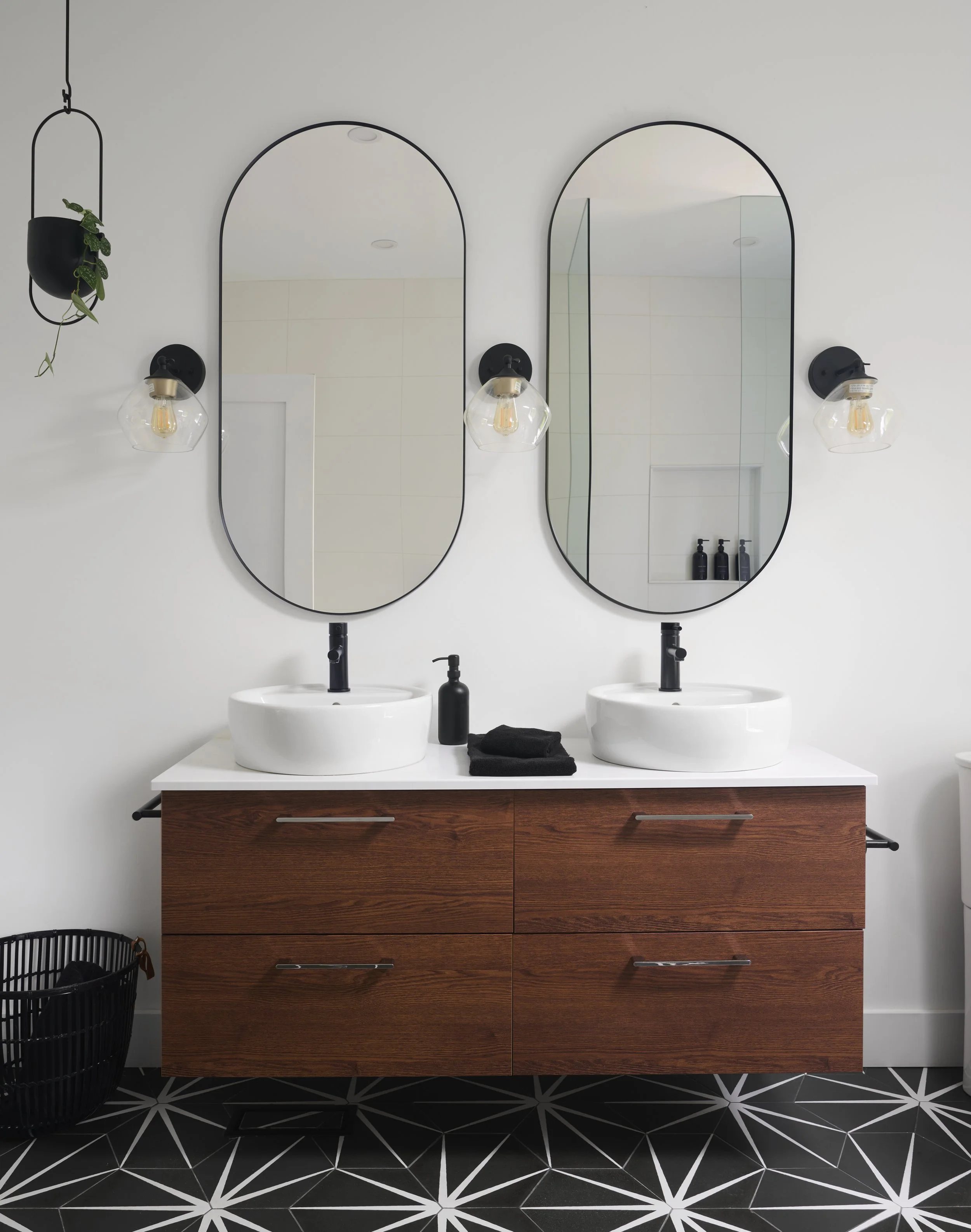 Modern bathroom with double vanity, two oval mirrors, black fixtures, wall-mounted lights, wooden cabinet, and black and white geometric tile floor.