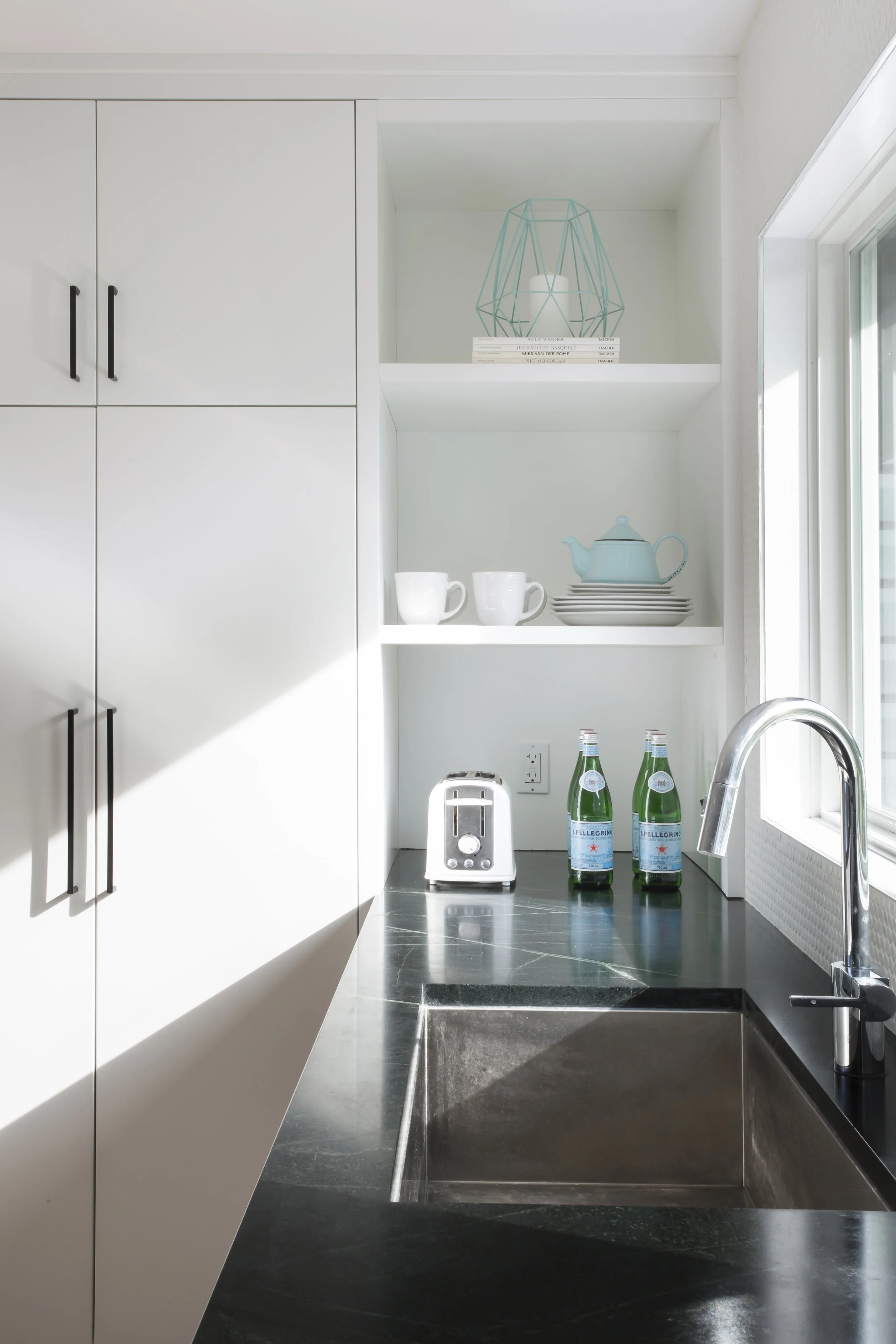 Modern kitchen with white cabinets, a black countertop, and a stainless steel sink next to a window. Shelving contains a decorative wire frame, books, mugs, a teapot, some plates, a toaster, and bottles of sparkling water.