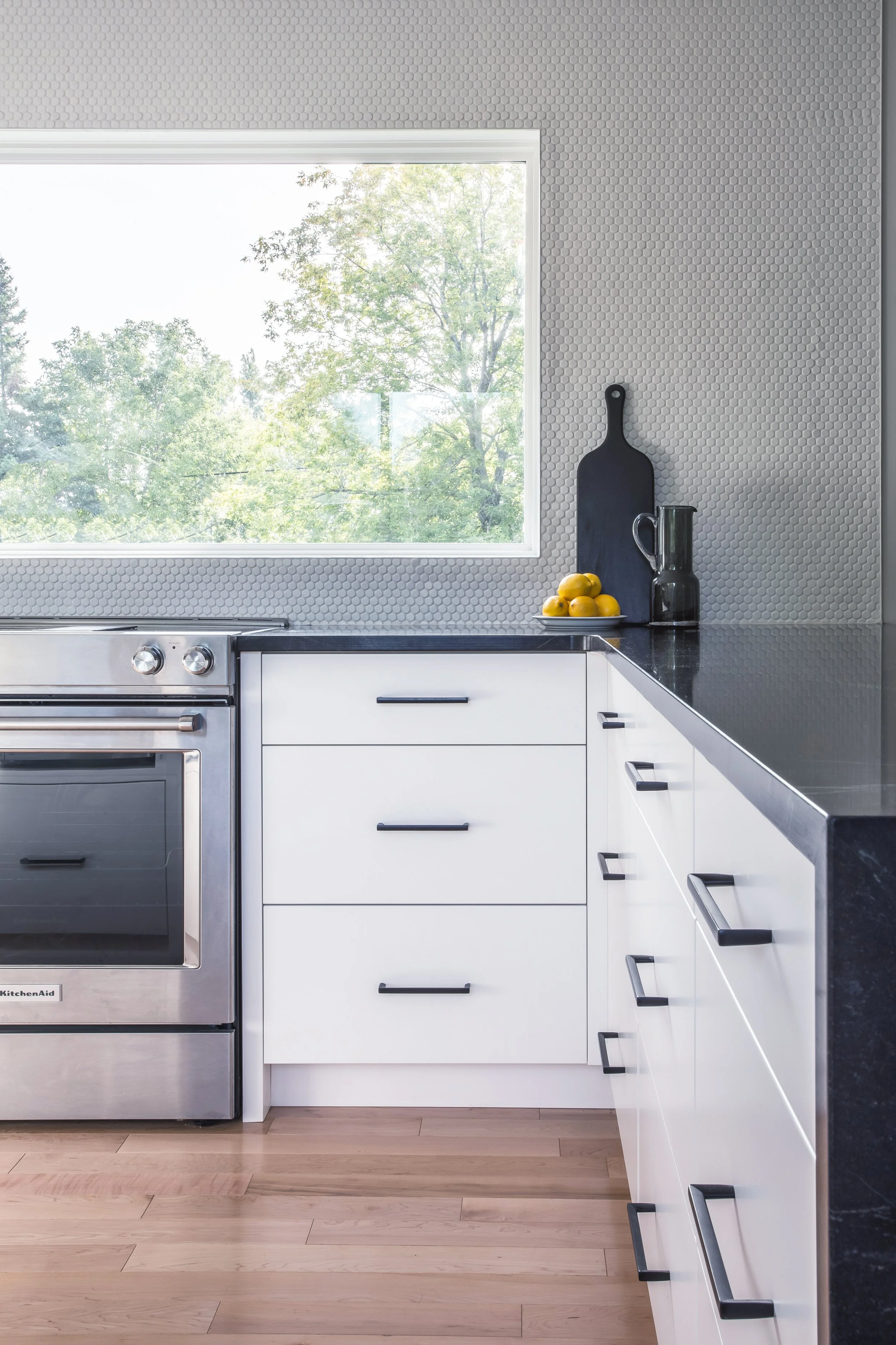 Modern kitchen with white cabinets, black handles, a black countertop, a window overlooking green trees, a cutting board, a vase, and lemons.
