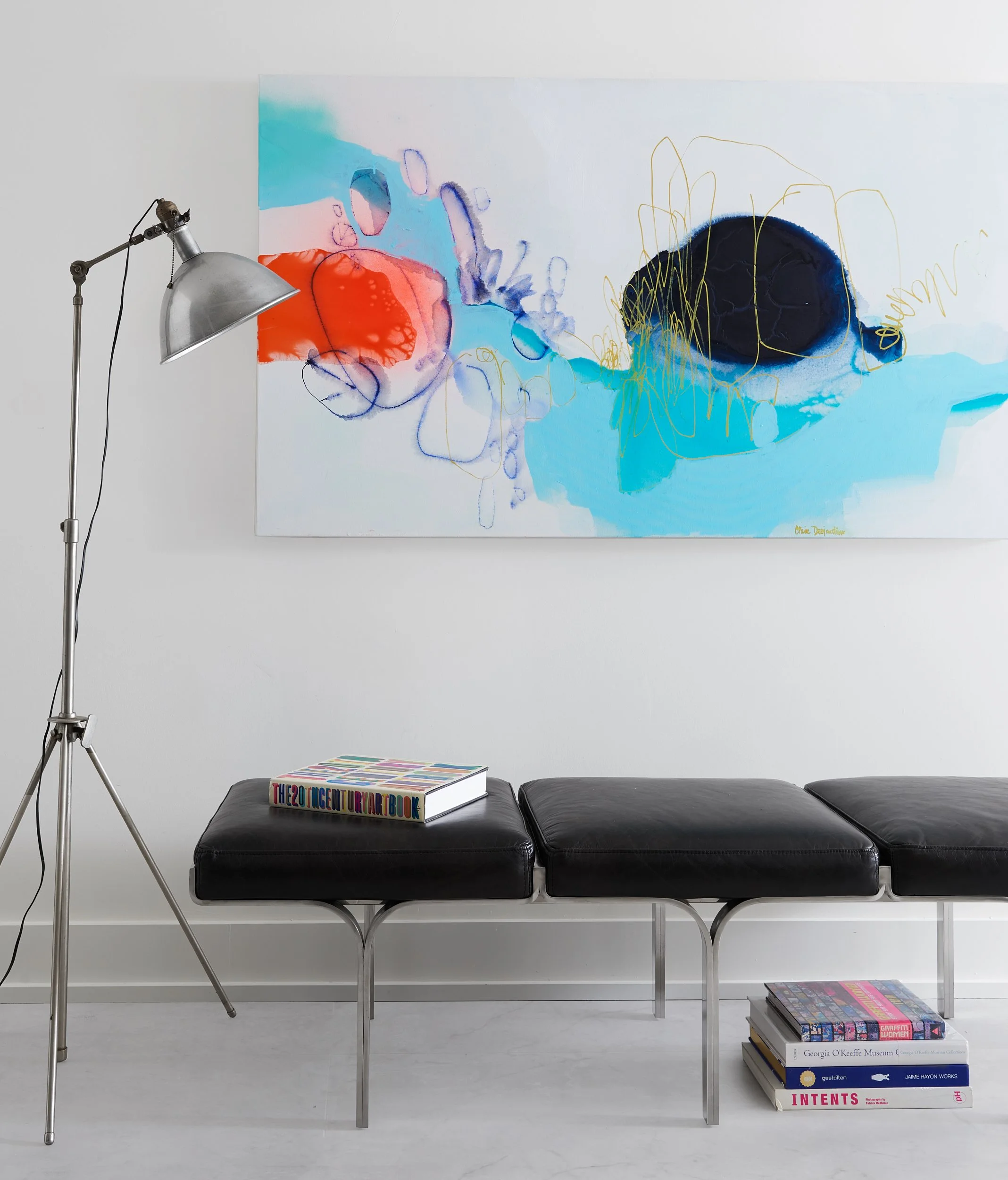 A modern living space featuring an abstract painting with blue, red, black, and yellow colors hung on a white wall, a black leather bench with a colorful art book on top, and a stack of art books underneath. A tall adjustable metal floor lamp stands 