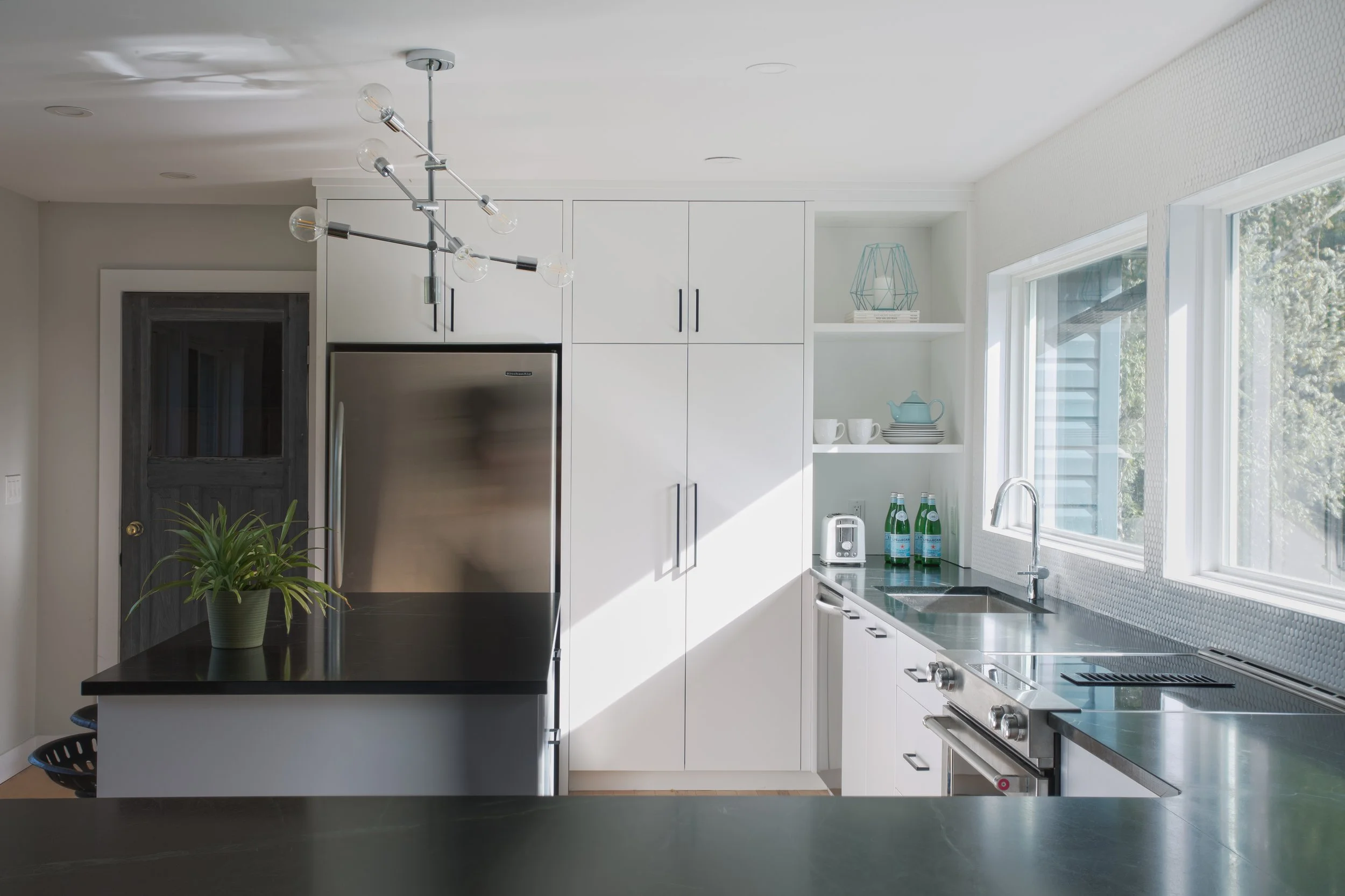 Modern kitchen with white cabinets, black countertops, stainless steel appliances, open shelving with dishes and teapot, large window, and a black island with a potted plant.