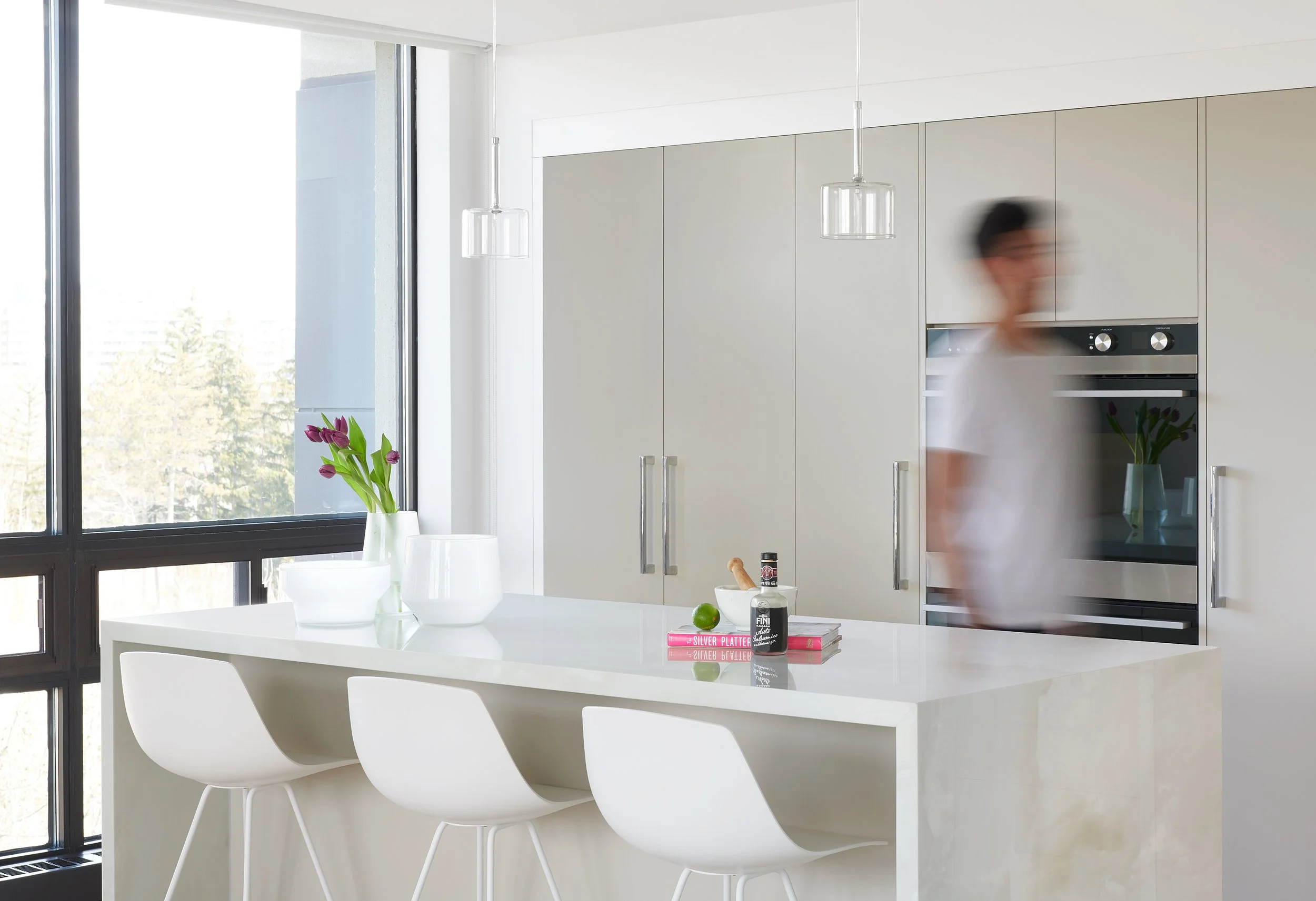 Modern kitchen with a large white island, white chairs, a vase of purple tulips, and built-in appliances. A person is blurred in motion near the cabinets.