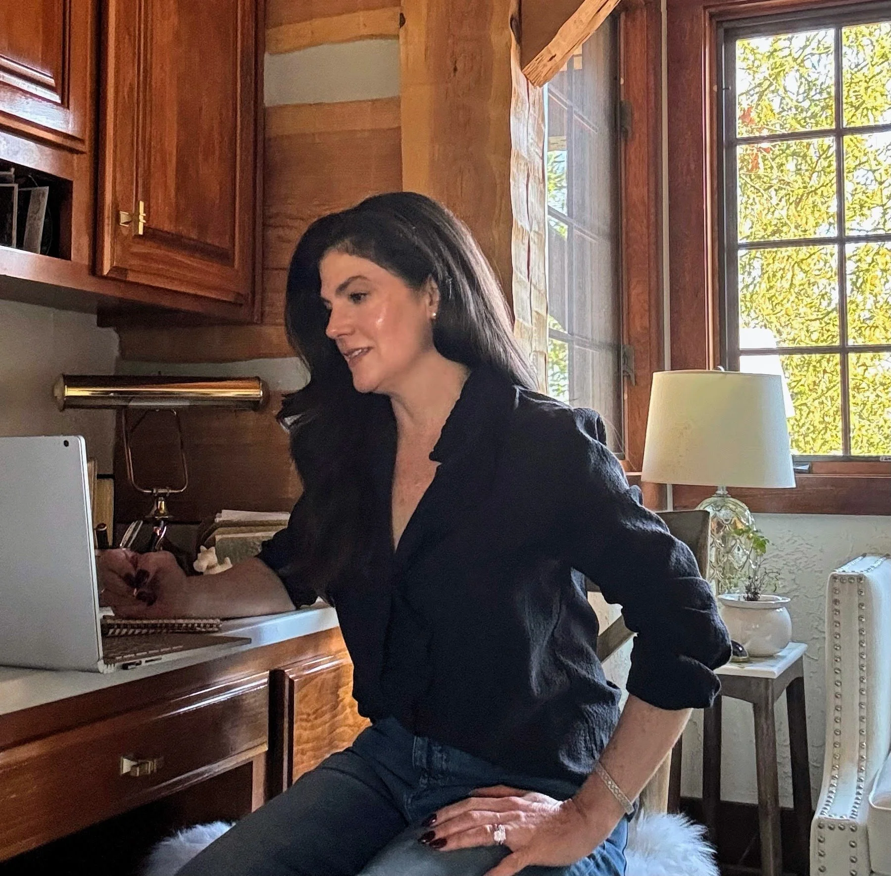 Woman with long dark hair sitting at a desk working on a laptop in a cozy room with wooden walls and large windows letting in natural light.