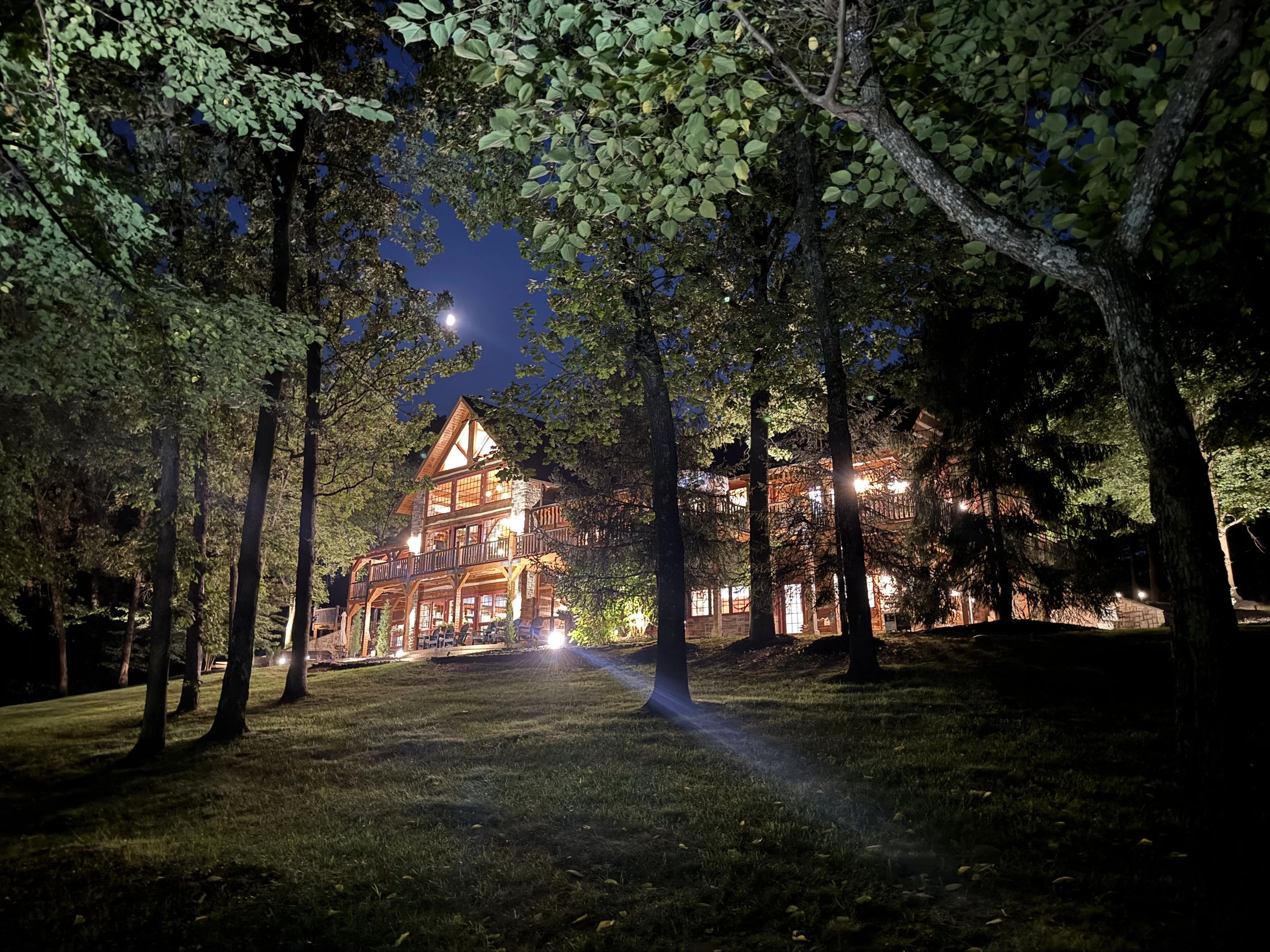 Night view of a well-lit, large wooden house surrounded by trees and a grassy yard, with moon visible in the sky.