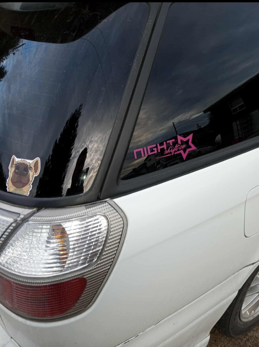 The rear window of a white subaru jdm car with a pink and black sticker that says 'NIGHTSHIFT.CO' with a star design in the upper right corner.