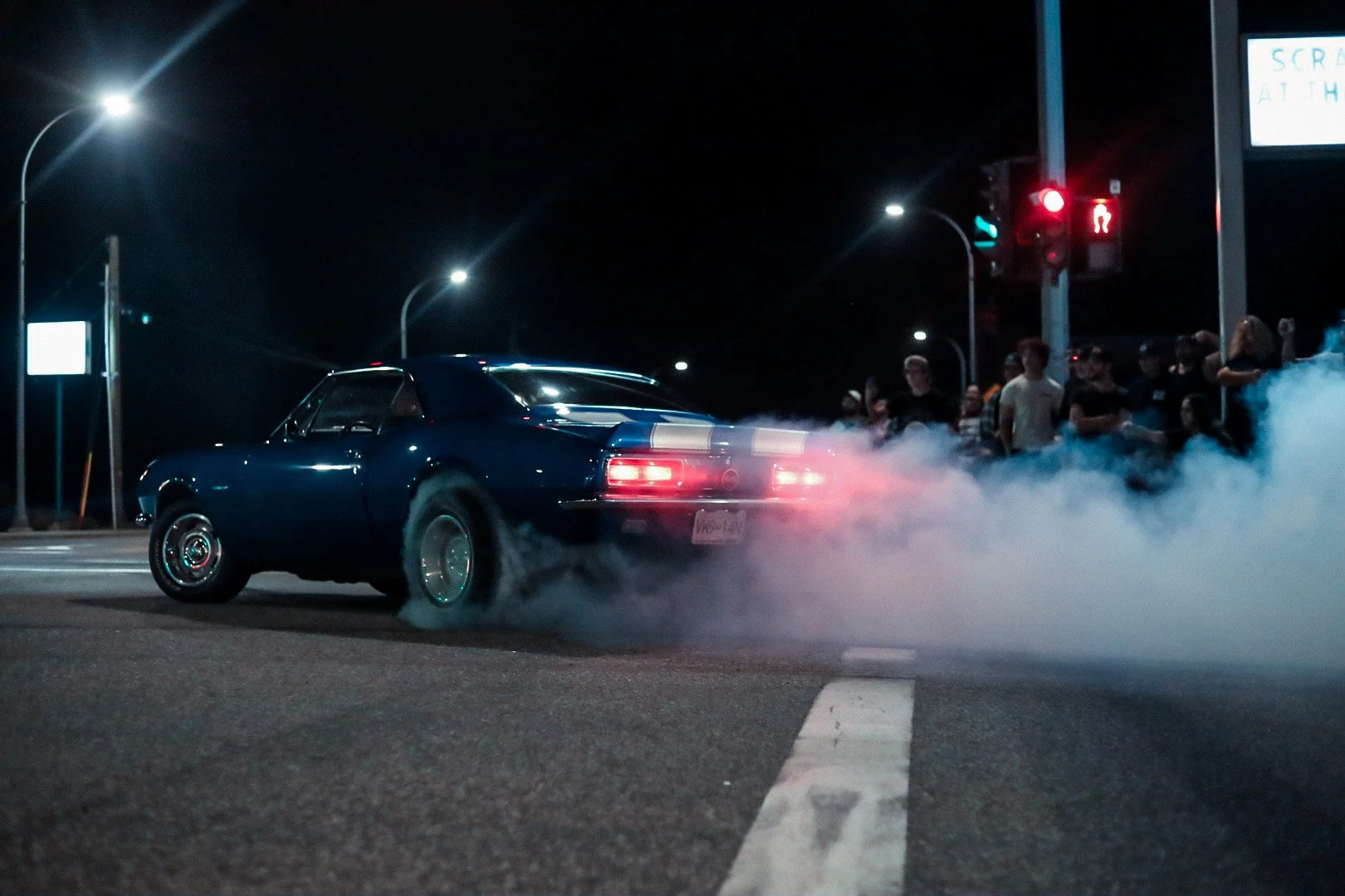 A classic blue sports car performing a burnout on a city street at night with smoke coming from the tires, and people gathered on the sidewalk watching.