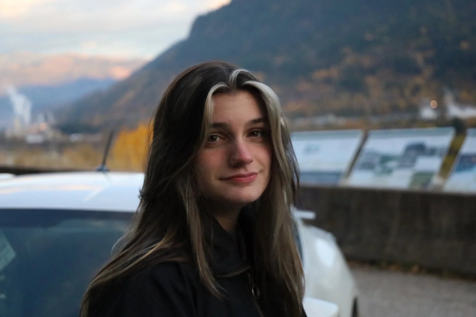 A young woman with long dark hair and light streaks, smiling slightly, is outdoors with mountains and a partly cloudy sky in the background.