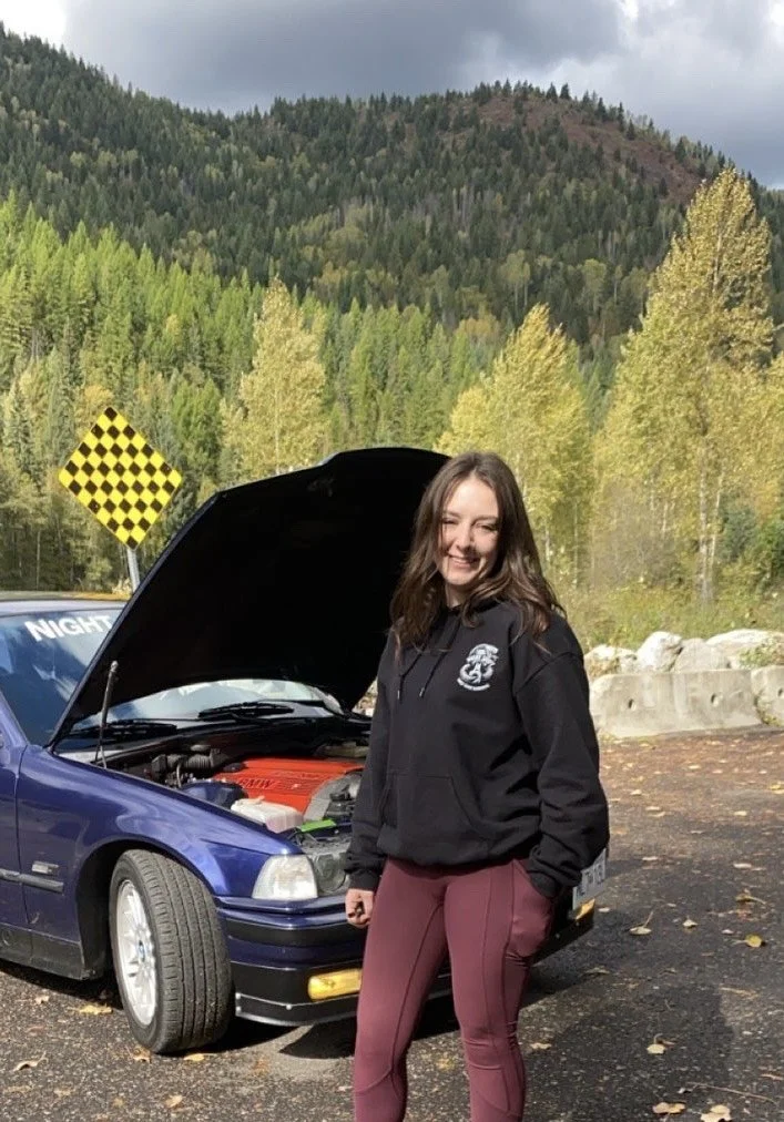 A woman with brown hair smiling and standing in front of a blue car with the hood open, in a scenic outdoor area with trees and mountains in the background.