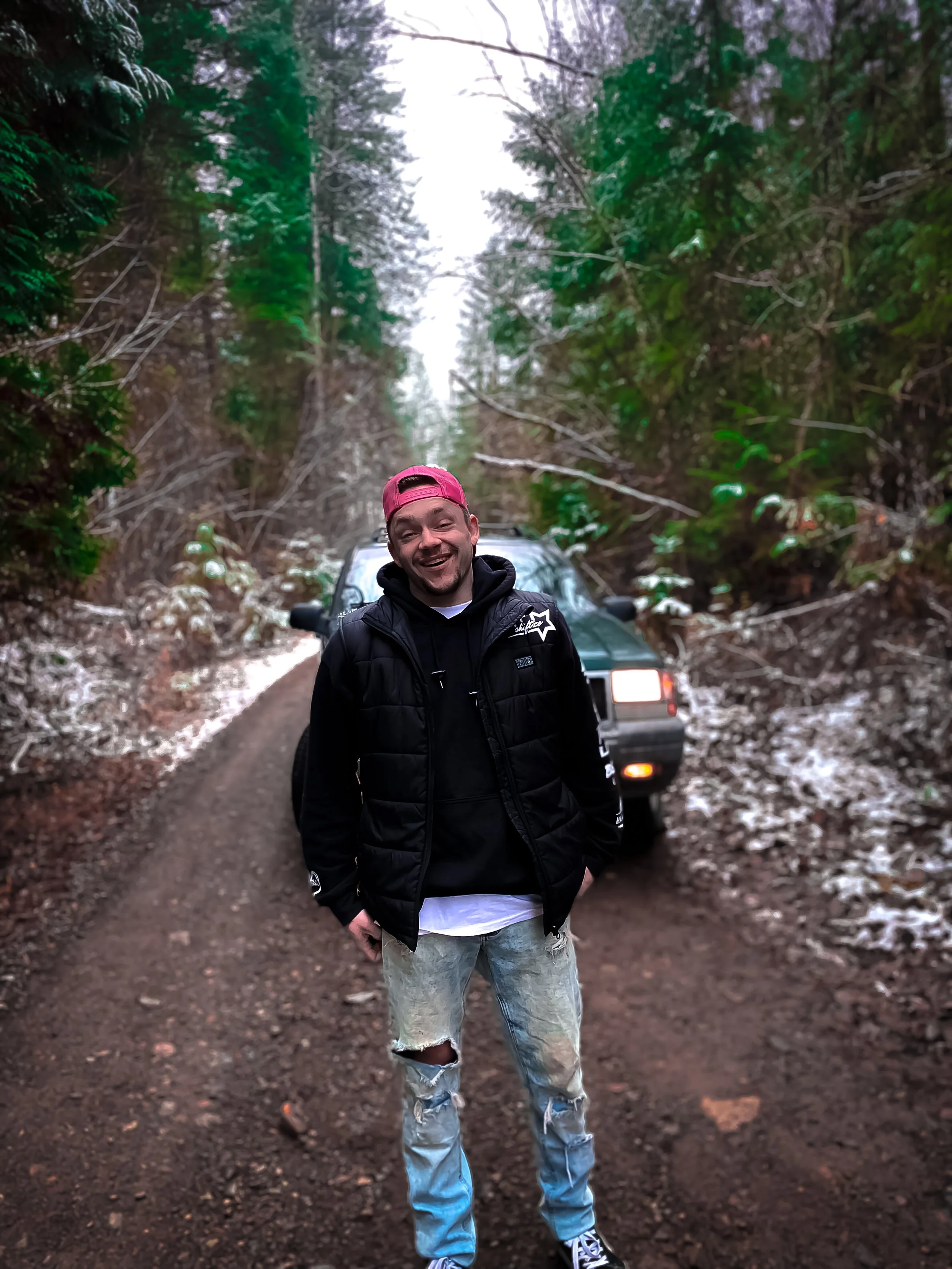 A young man in a black jacket and ripped jeans standing on a dirt forest road, smiling at the camera, with tall trees and a vehicle behind him.