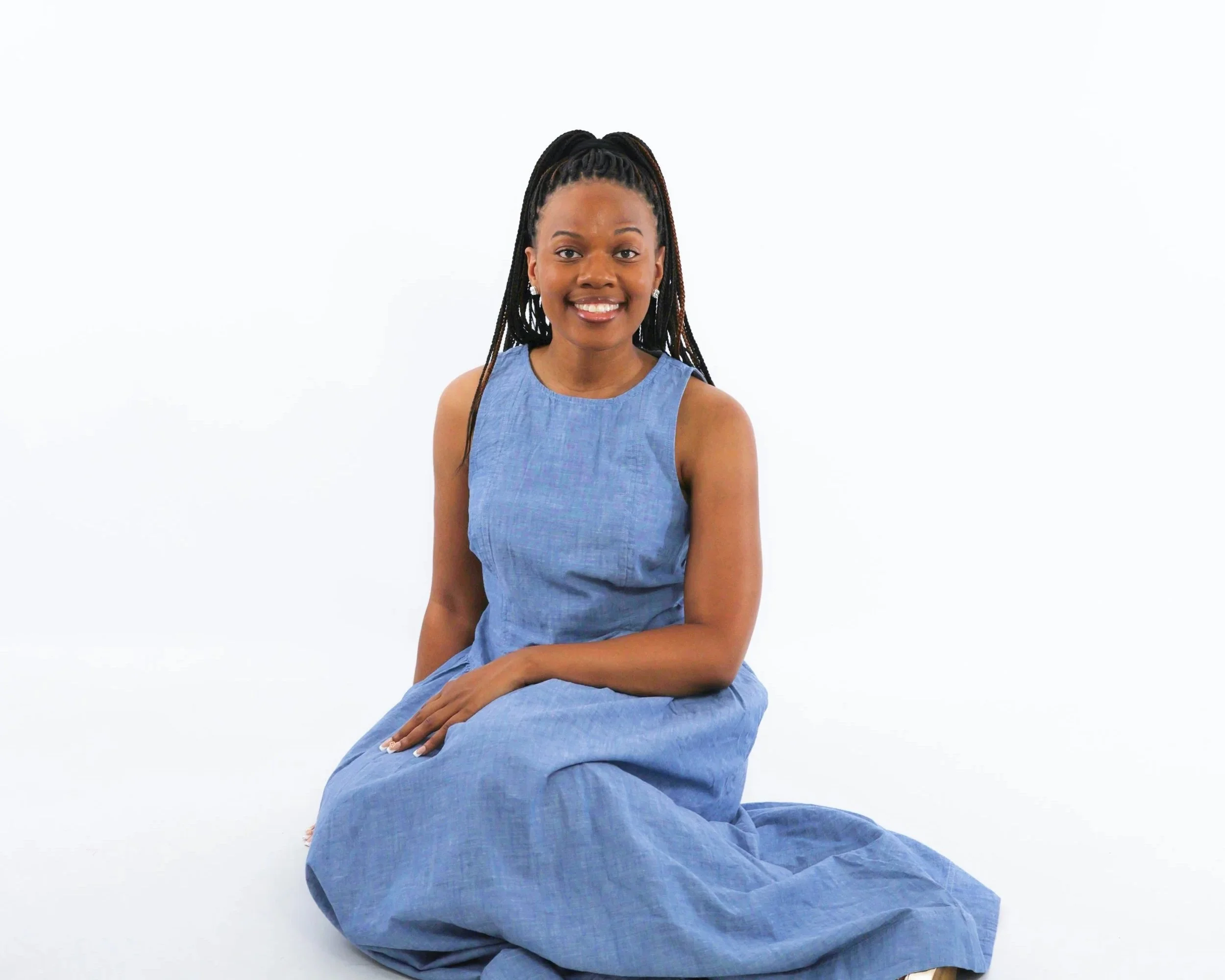 A woman with braided hair wearing a blue sleeveless dress, sitting on a large blue cloth, against a plain white background.