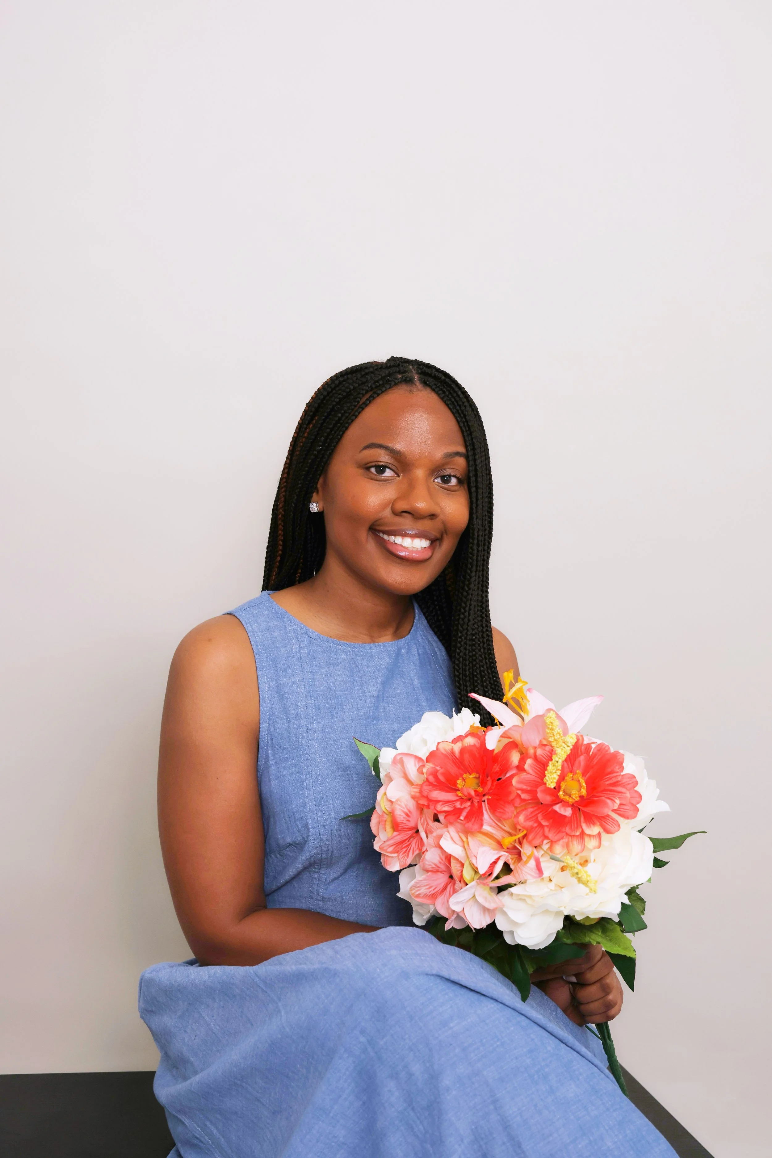 A young woman with long braided hair, wearing a sleeveless blue dress, holding a bouquet of pink and white flowers, smiling, sitting against a plain white wall.
