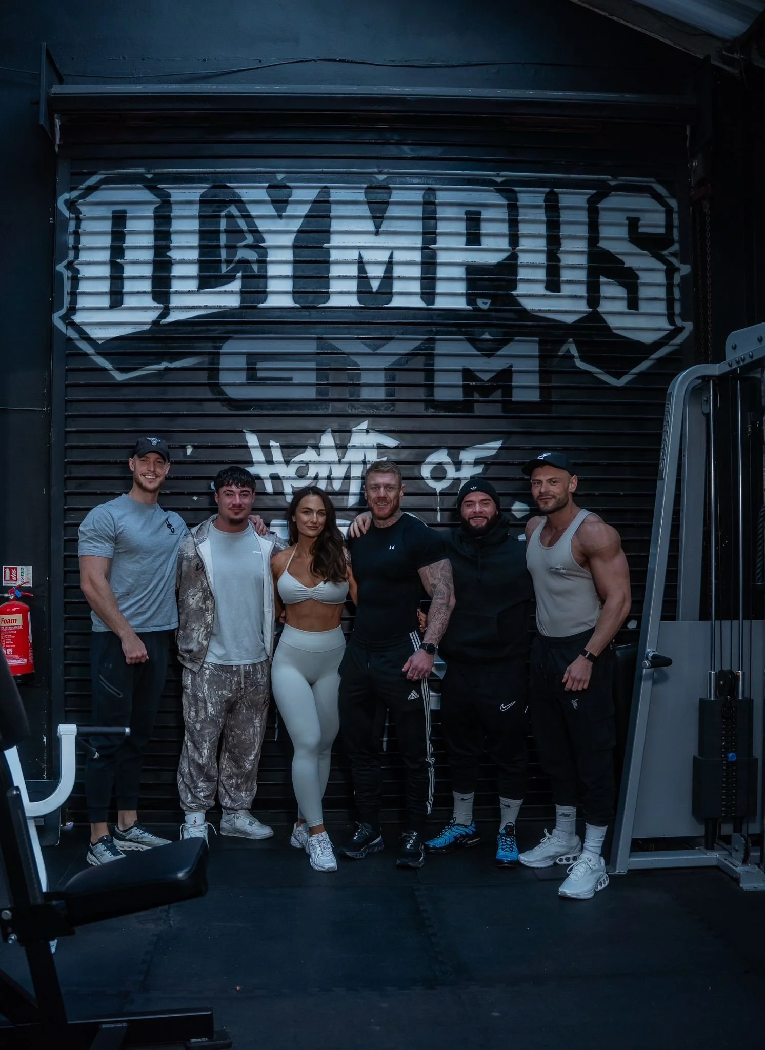Group of six fit people, including men and a woman, standing in gym in front of a large black and gray sign that reads "Olympus Gym" with graffiti-style text "Home of" below.