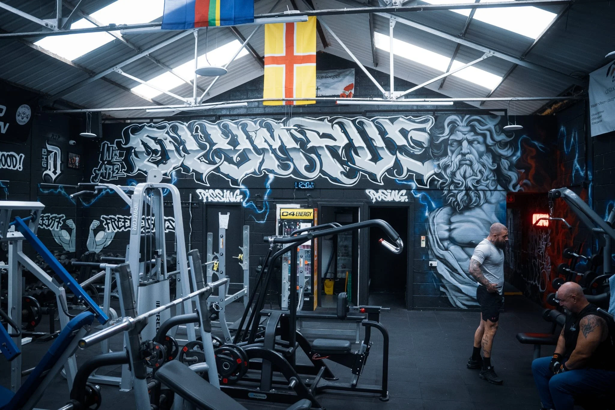 Inside a gym with black walls and graffiti art, including a large face of a bearded man, fitness equipment like weights and machines, and two men, one standing and one sitting.