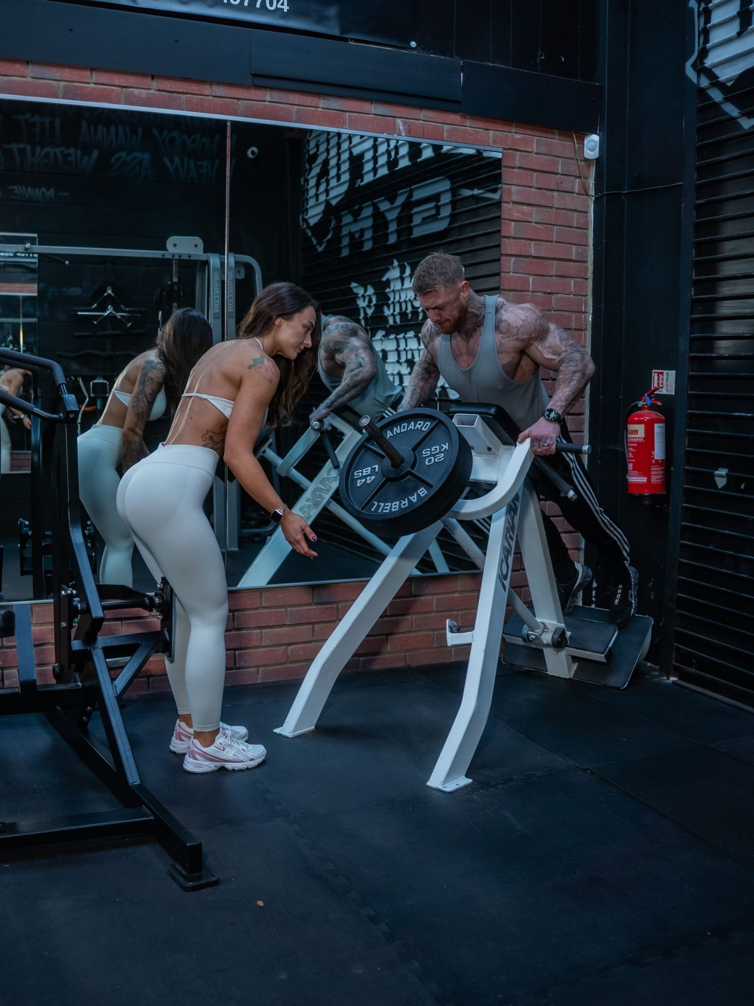 Two women and two men working out together at a gym. One woman is adjusting weights on a machine while the other woman and a man are lifting weights on a different machine. The gym has brick walls and large mirrors.