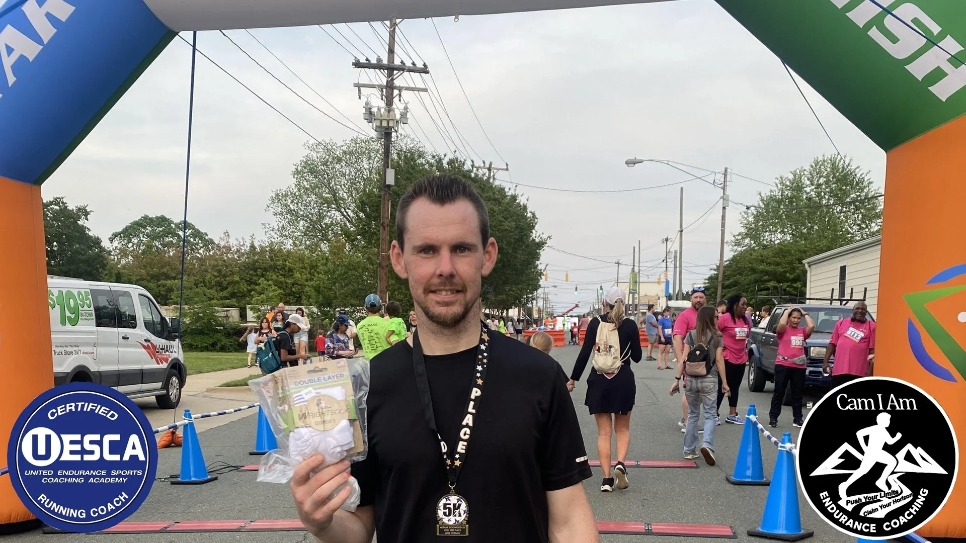 A man holding a gift bag at a race event with a crowd of runners and spectators behind him. There are blue cones and banners with logos, including UESCA and 'Cam I Am Endurance Coaching,' at the finish line.