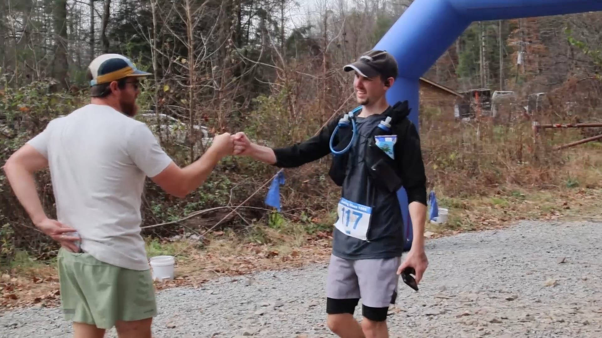 Two men shaking hands at a race event near a wooded area, with a blue inflatable arch and gravel path in the background.
