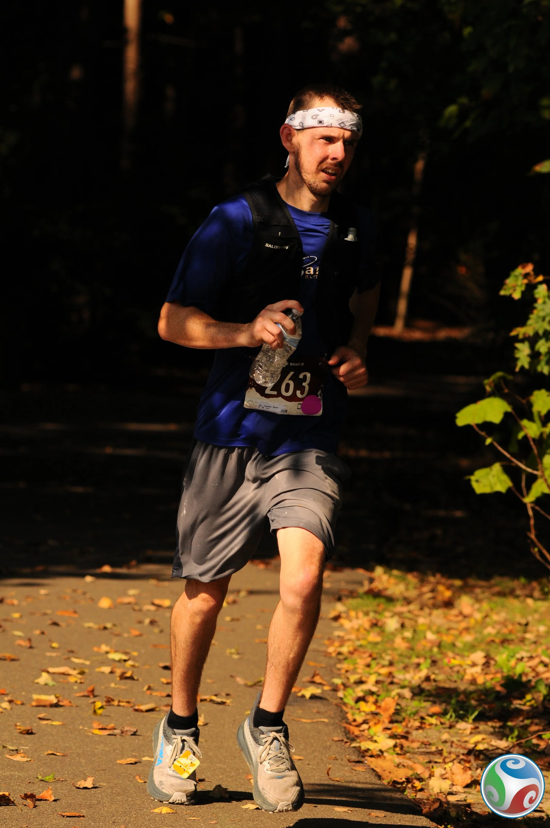 A man participating in a marathon, wearing a race bib numbered 63, a white headband, a blue shirt, gray shorts, gray running shoes, and carrying a water bottle, running along a leaf-covered path in a wooded area.