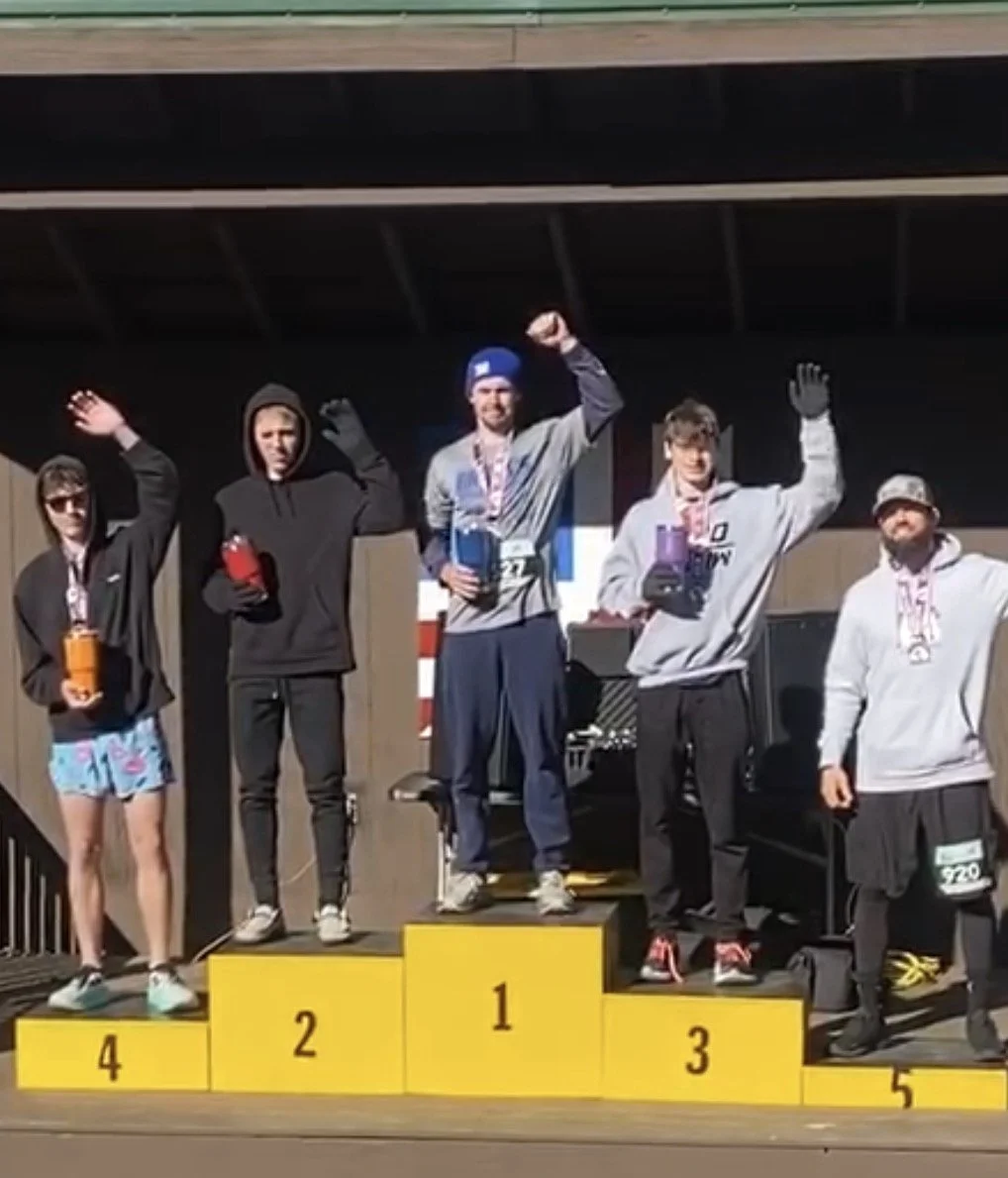 Podium award ceremony with five male athletes holding drinks and medals, standing on first, second, third, fourth, and fifth place steps, outdoors under a wooden structure.
