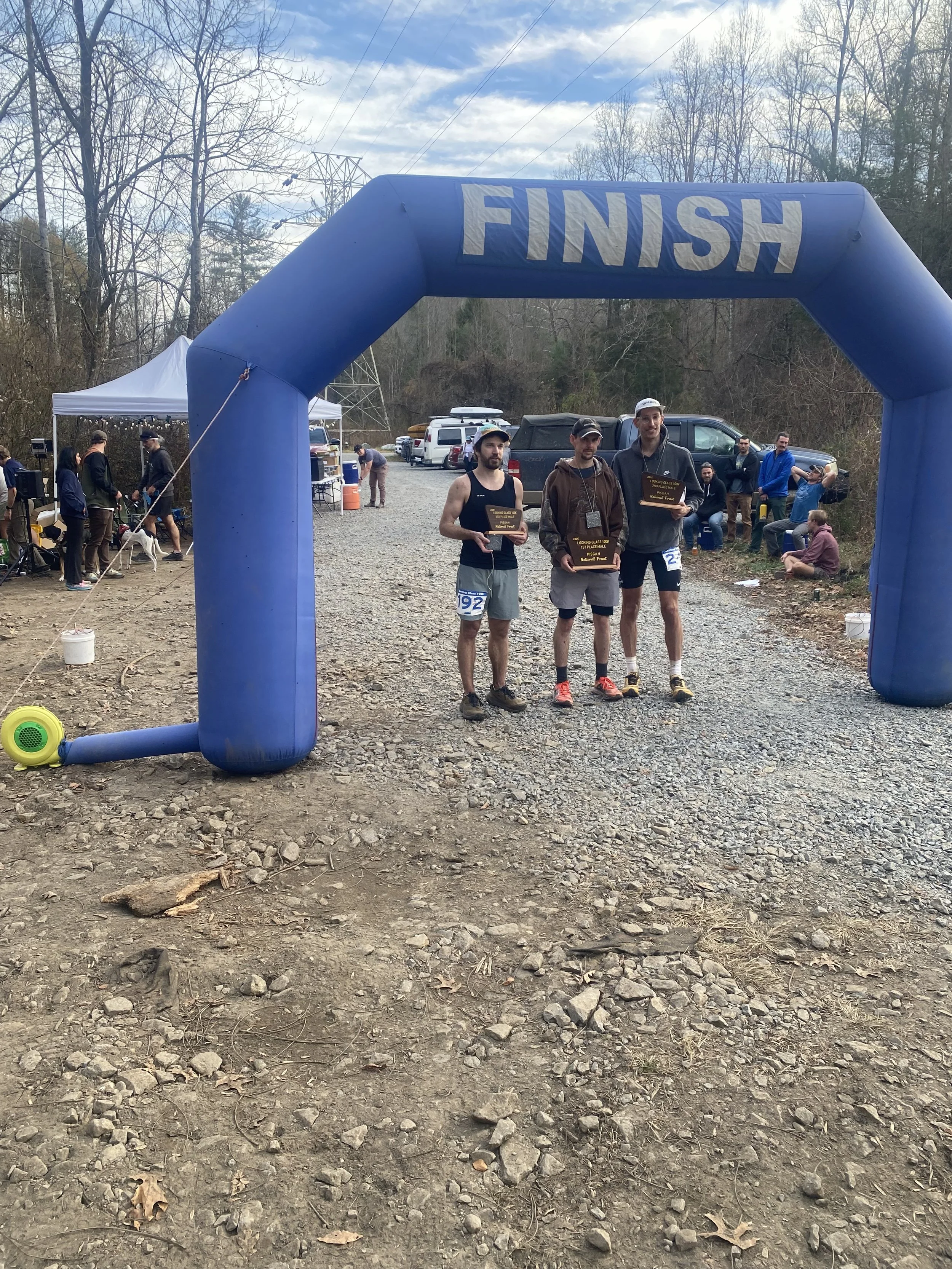 Three male runners standing under a blue inflatable finish line arch, holding plaques, at an outdoor race event on a dirt trail with trees and spectators in the background.