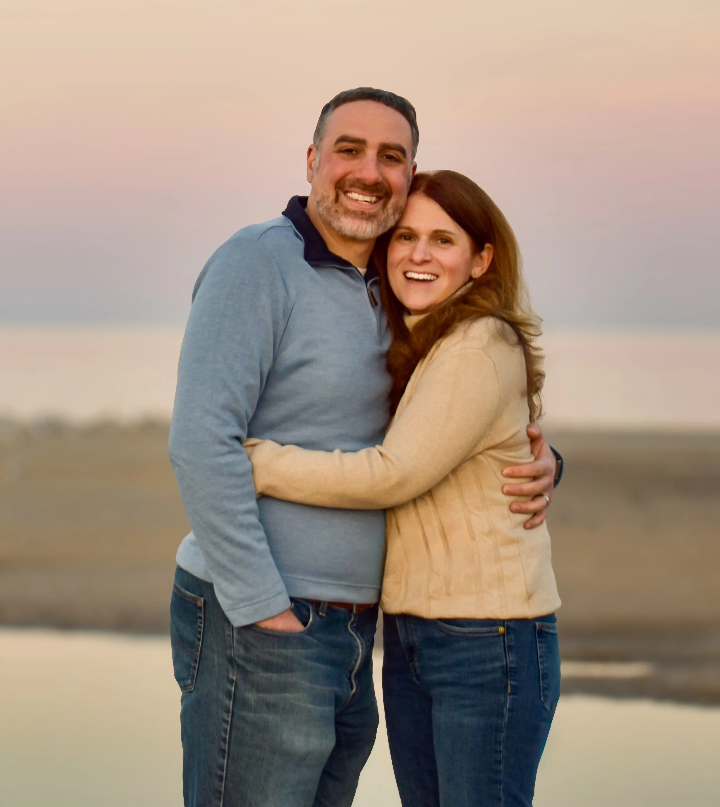 A happy couple embracing on the beach during sunset, smiling at the camera.