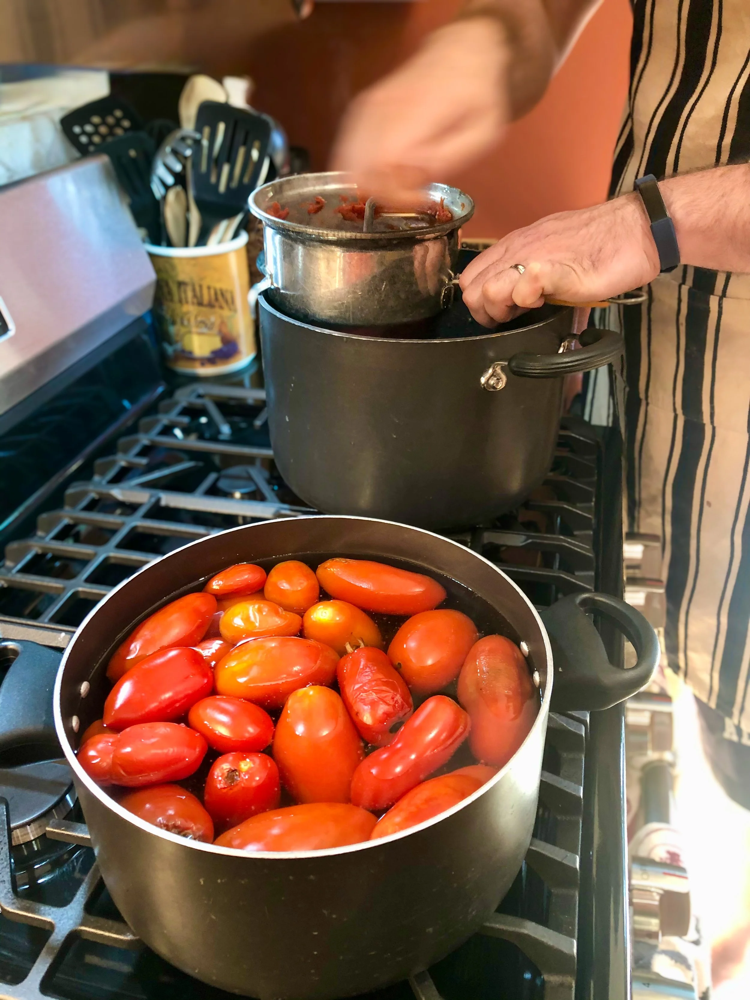 Two pots on a stove, one with plum tomatoes and the other with a pot of tomato sauce being stirred. Making homemade tomato sauce from garden grown tomatoes.