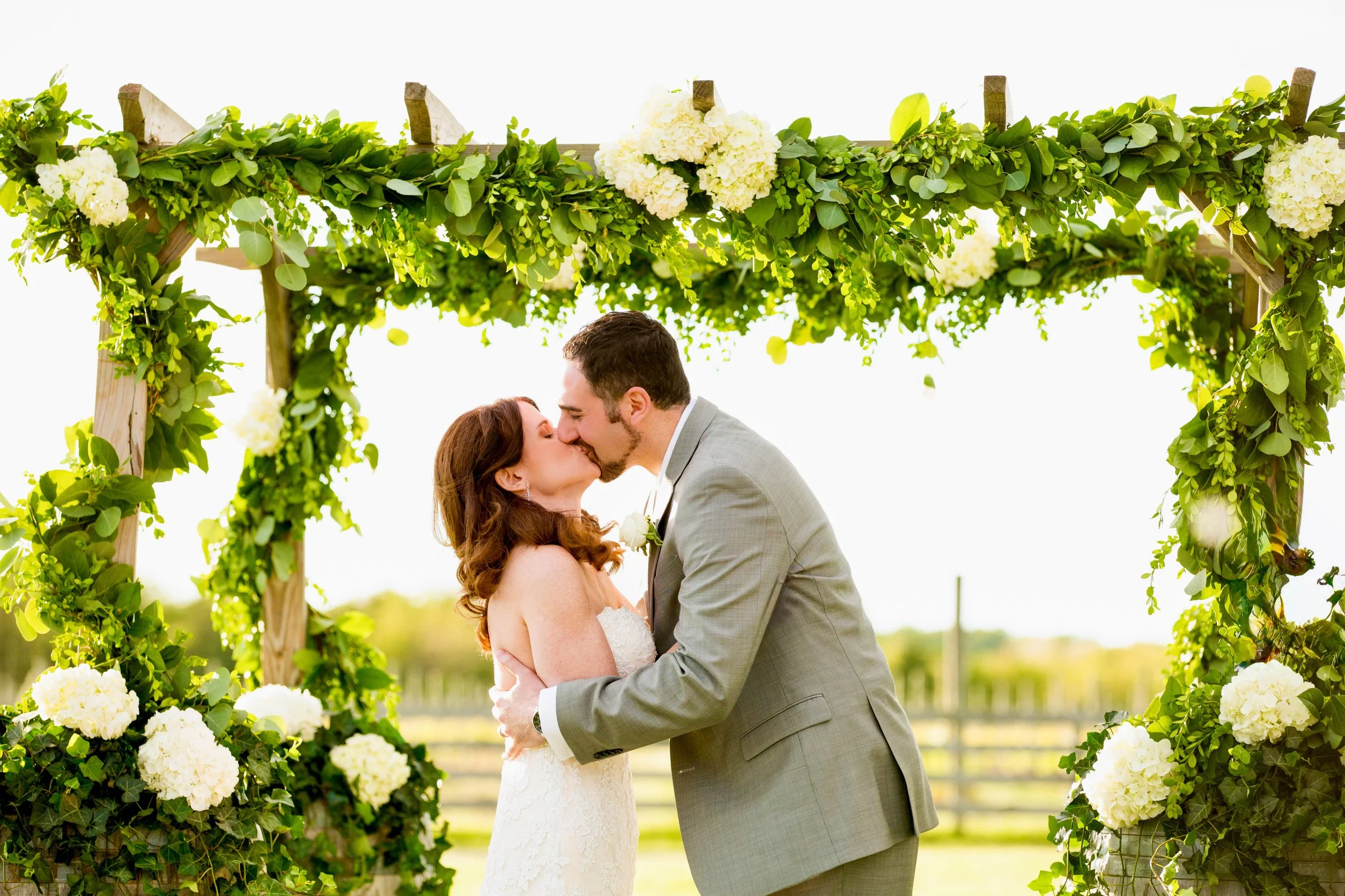 A bride and groom sharing a kiss under a decorated wedding arch made of green leaves and white flowers outdoors.