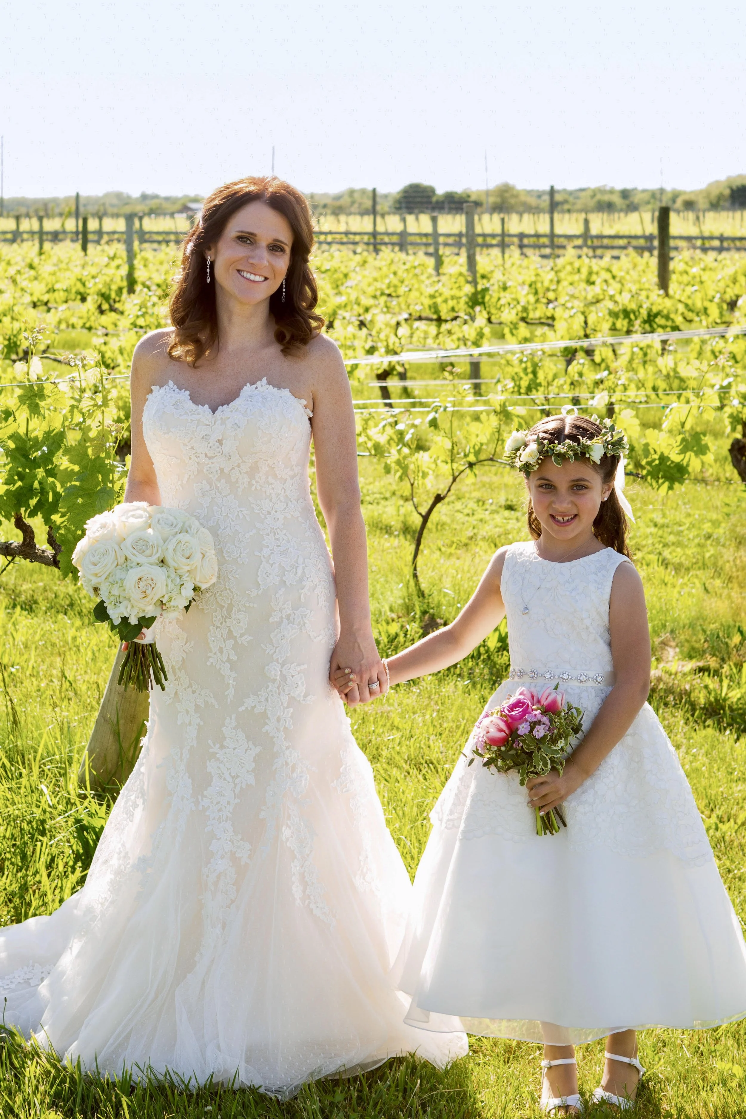A woman in a white wedding dress holding a bouquet, and a young girl in a white dress with a floral crown holding a small bouquet, standing hand-in-hand outdoors in front of a green vineyard under a bright sky.