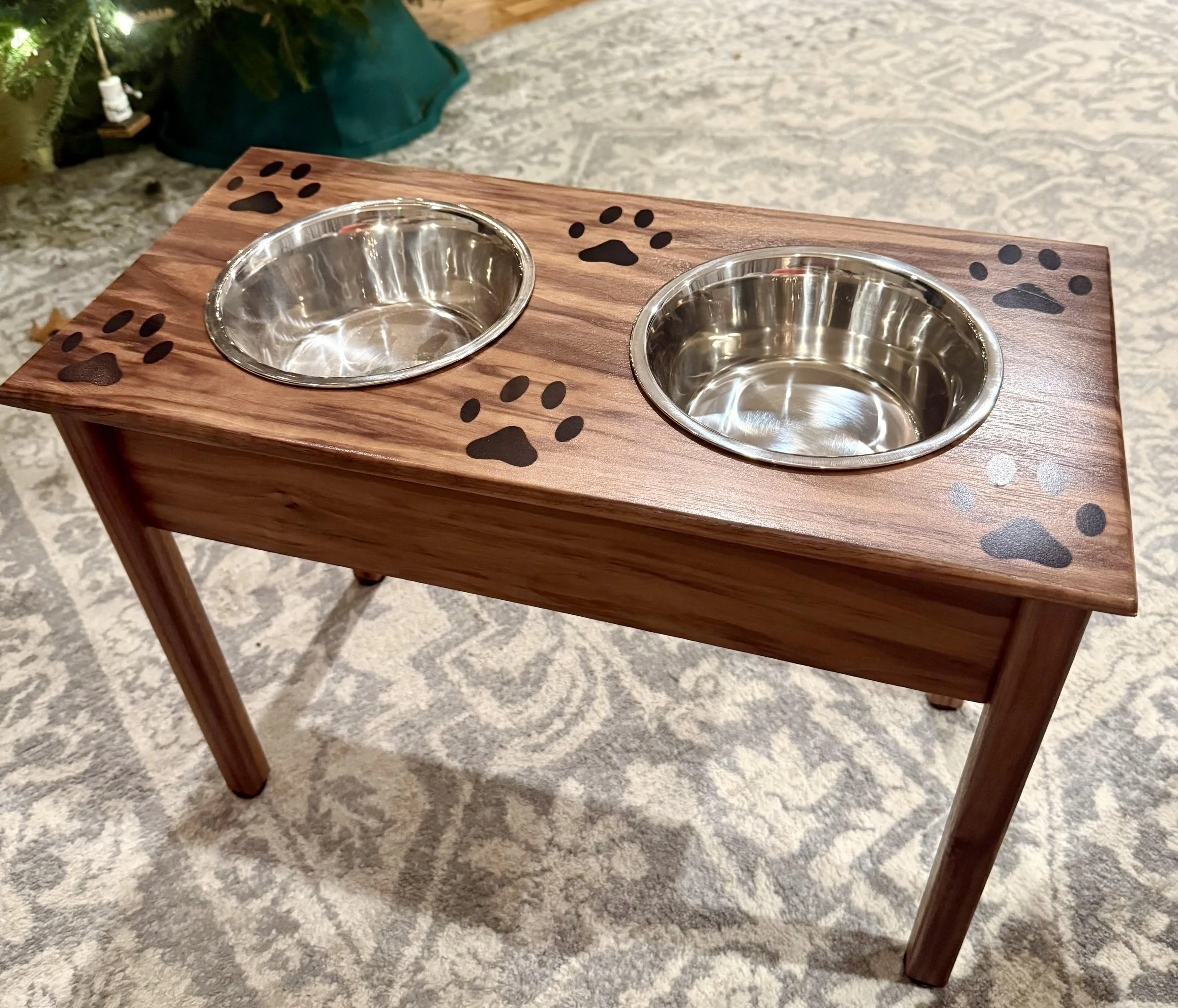 A wooden elevated dog feeding station with two stainless steel bowls, decorated with black paw prints, placed on a beige patterned carpet indoors.