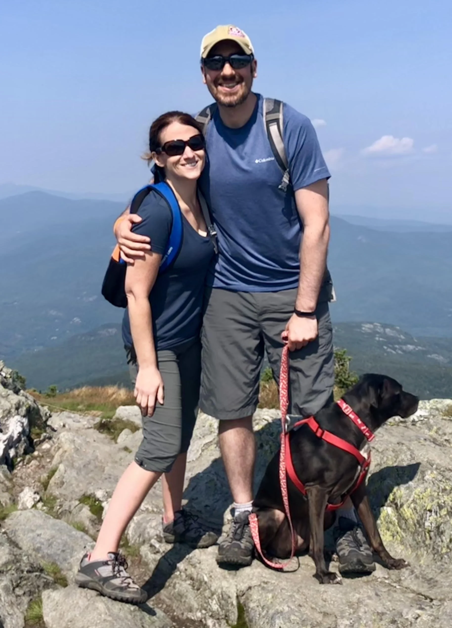 A smiling couple with a dog on a mountain hiking trail, with mountain scenery and blue sky in the background. Family looking to adopt a baby.